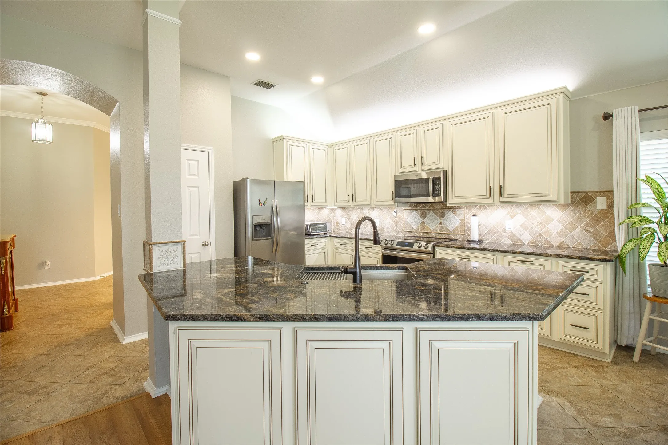 Kitchen featuring an island with a black countertop and an integrated sink, cream-colored cabinetry, stainless steel appliances, and mosaic tile backsplash