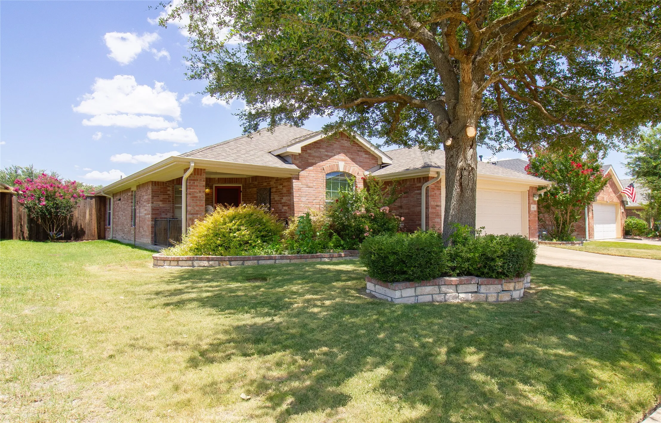Brick exterior residence featuring a prominent mature tree, landscaped beds with stone block edging, a covered front entry, and an attached garage with a white door