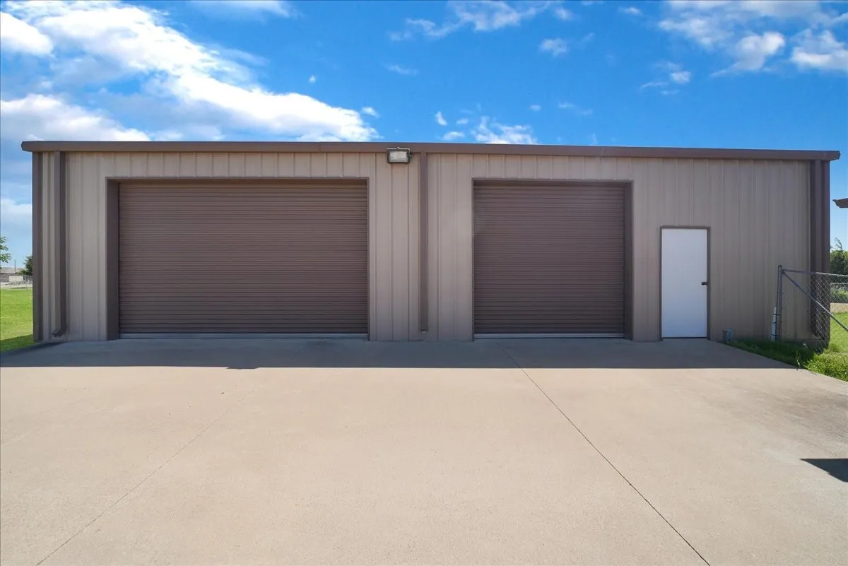Metal-sided structure featuring two large roll-up doors, a pedestrian entry door, an exterior light fixture, and a concrete approach