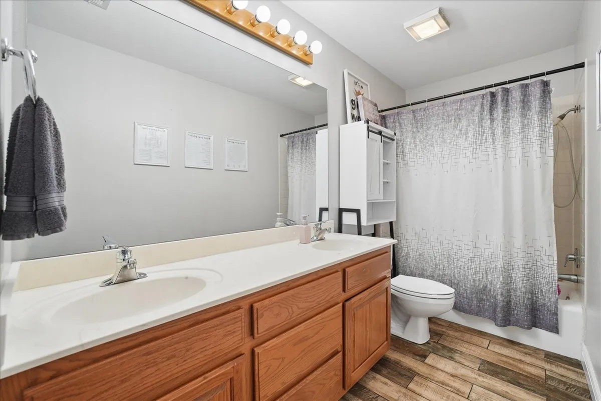 Bathroom featuring a dual-sink vanity with wood-finish cabinetry, a large mirror with overhead lighting, a built-in shower/tub combination, and wood-finish flooring