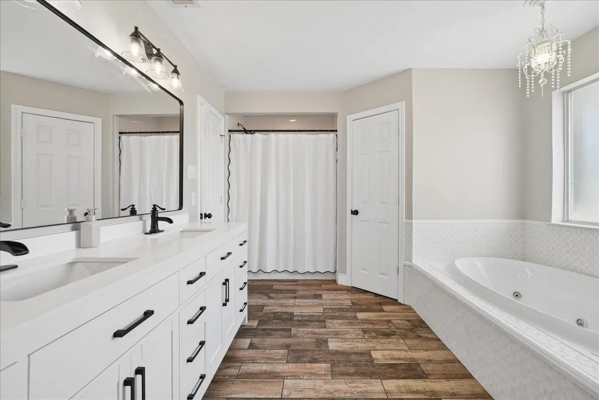 Spacious bathroom featuring a double vanity with white cabinetry and black hardware, quartz countertops, a large framed mirror, a separate shower enclosure, and a built-in jetted tub with patterned tile surround