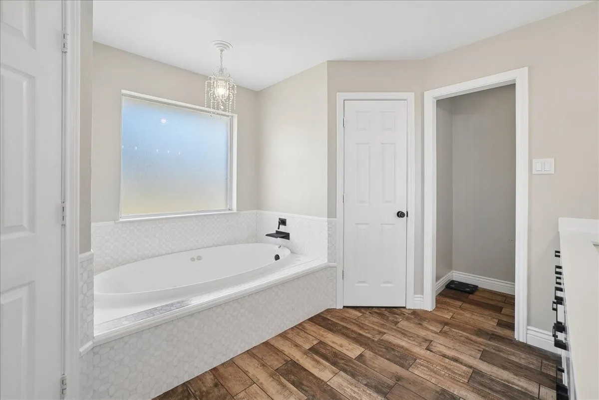 Spacious soaking tub with hex tile surround, frosted window, and a decorative chandelier