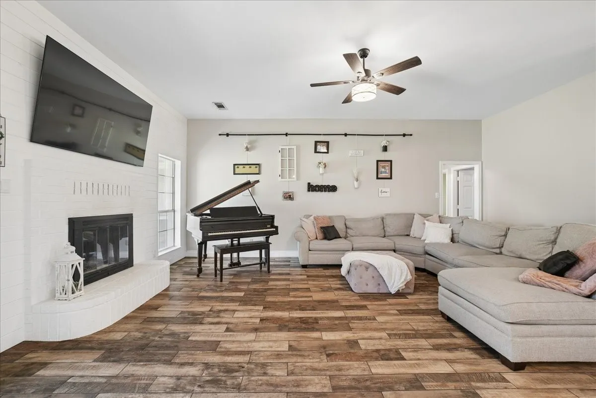 Spacious living area featuring wood-finish flooring, a white brick fireplace with a hearth, and a ceiling fan with integrated lighting