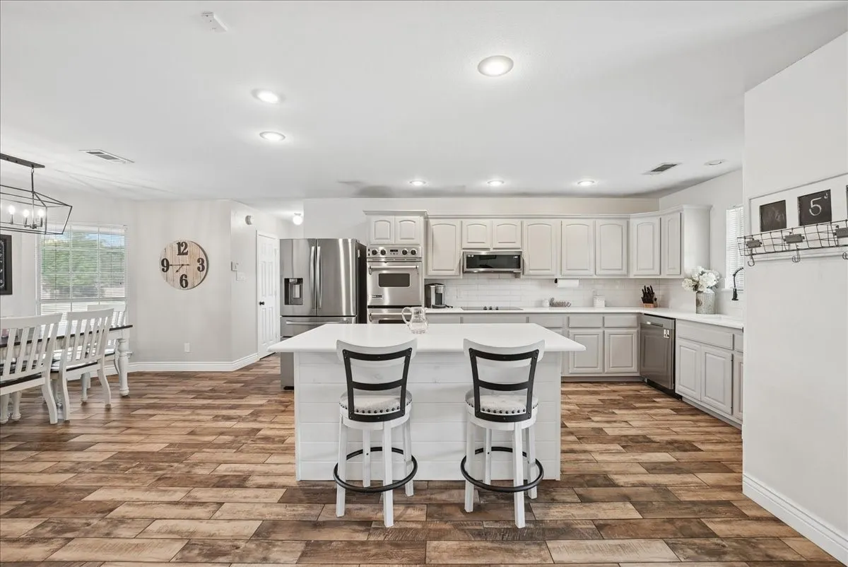 Open-concept kitchen featuring an expansive center island with seating, extensive cabinetry with white countertops, stainless steel appliances, and wood-finish flooring