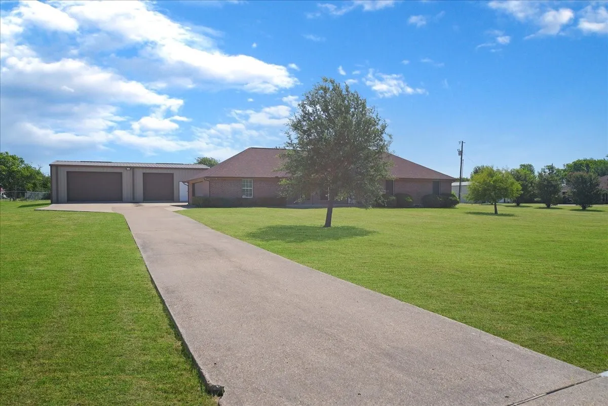 Expansive concrete driveway leading to a brick residence with a large detached metal garage featuring two bay doors