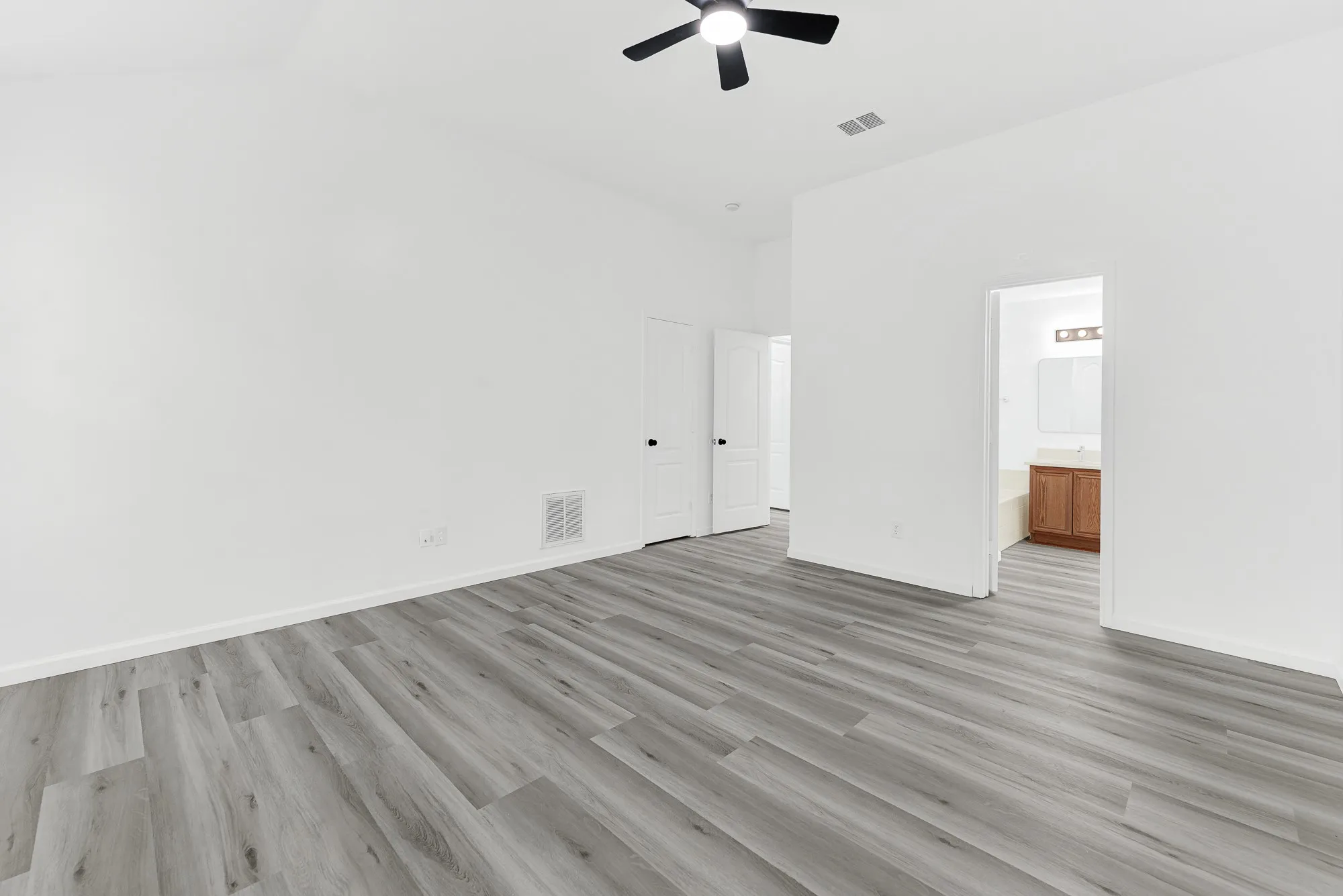 Spacious room with wood-finish flooring, crisp white walls, a ceiling fan, and direct access to a bathroom featuring a vanity with a light wood-tone cabinet and a mirror