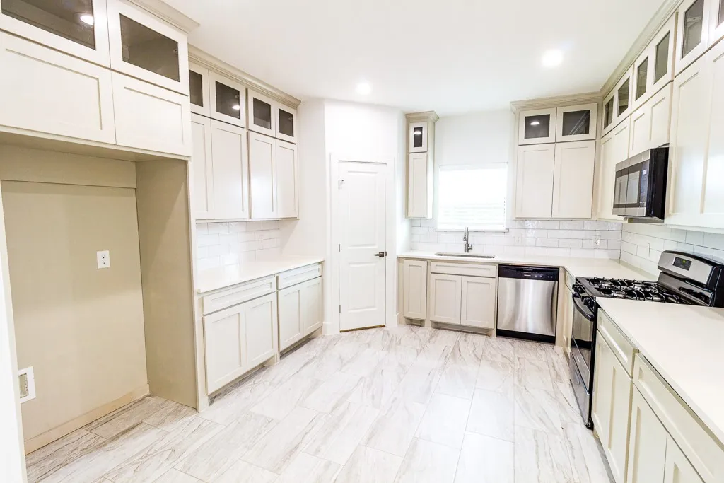 Bright, open kitchen featuring cream cabinetry with glass-front upper cabinets, stainless steel appliances including a gas range and dishwasher, white subway tile backsplash, light wood-finish flooring, and ample counter workspace with a center island