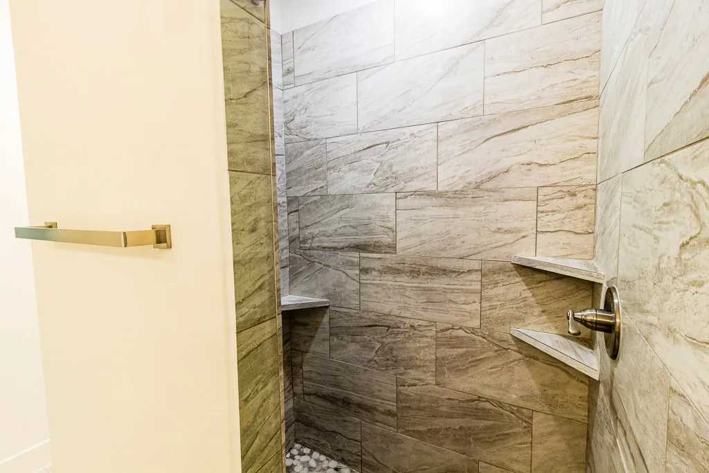 Tiled shower stall with an integrated bench, dual recessed shelving, brushed nickel fixtures, and a pebble-style floor