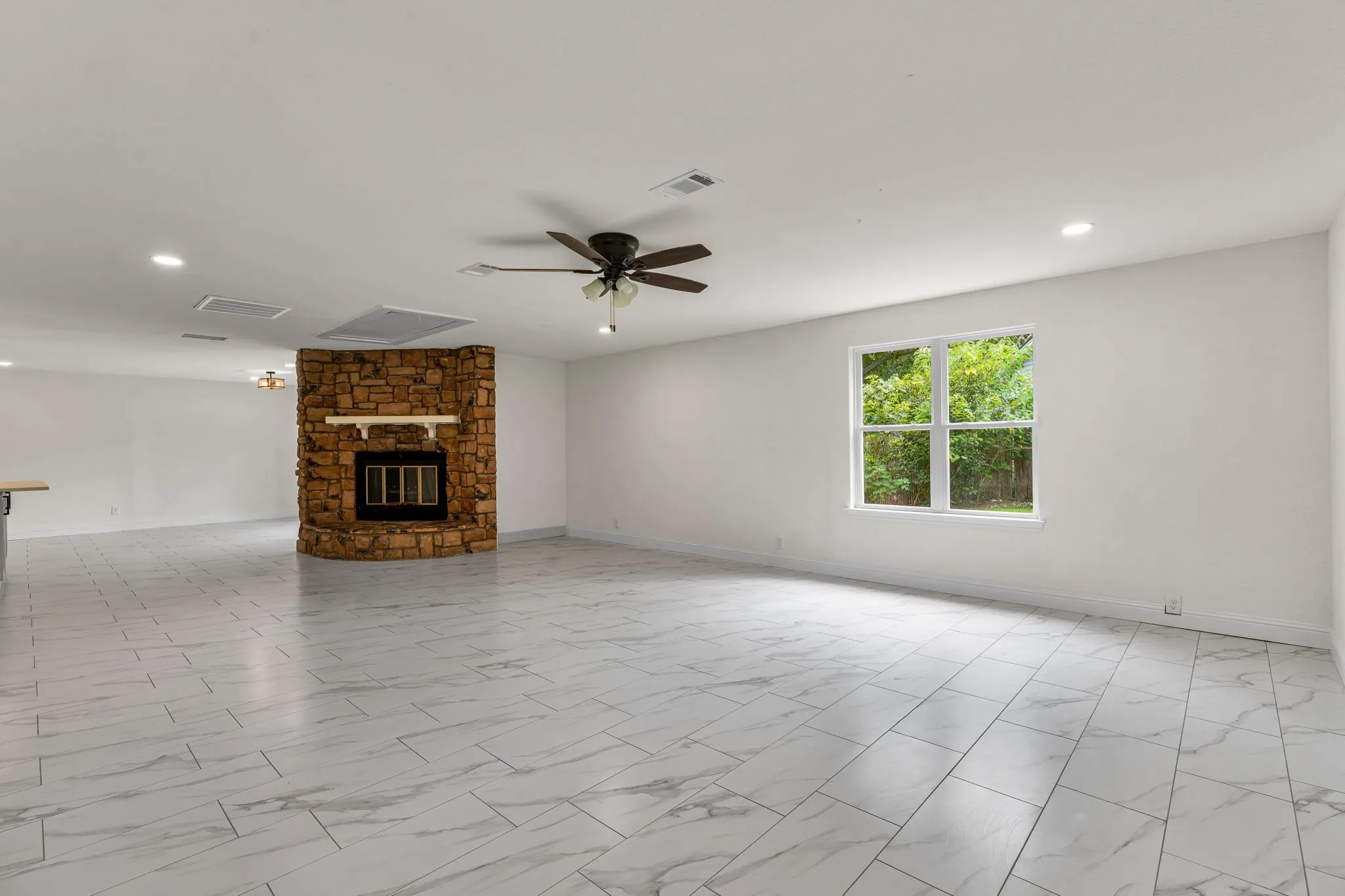 Spacious living area featuring a floor-to-ceiling stone fireplace with a wood mantle, large window, ceiling fan, recessed lighting, and marble-look tile flooring