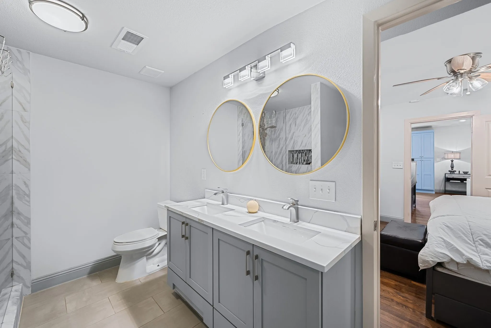 Bathroom featuring a double vanity with light gray cabinetry and white countertops, dual circular gold-framed mirrors, and a herringbone-patterned shower surround