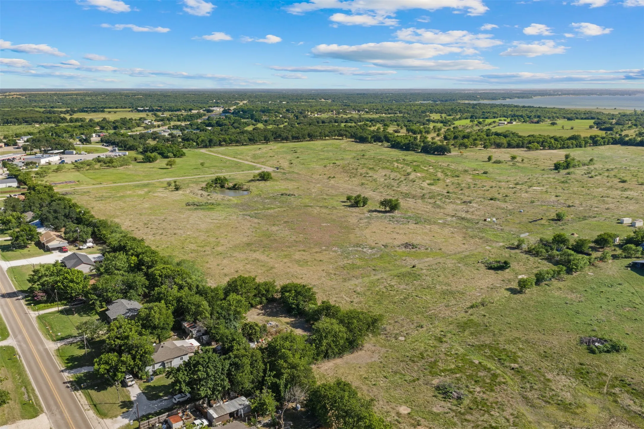 Expansive open land featuring a mix of green fields and scattered trees, bordered by a paved road and existing residential structures