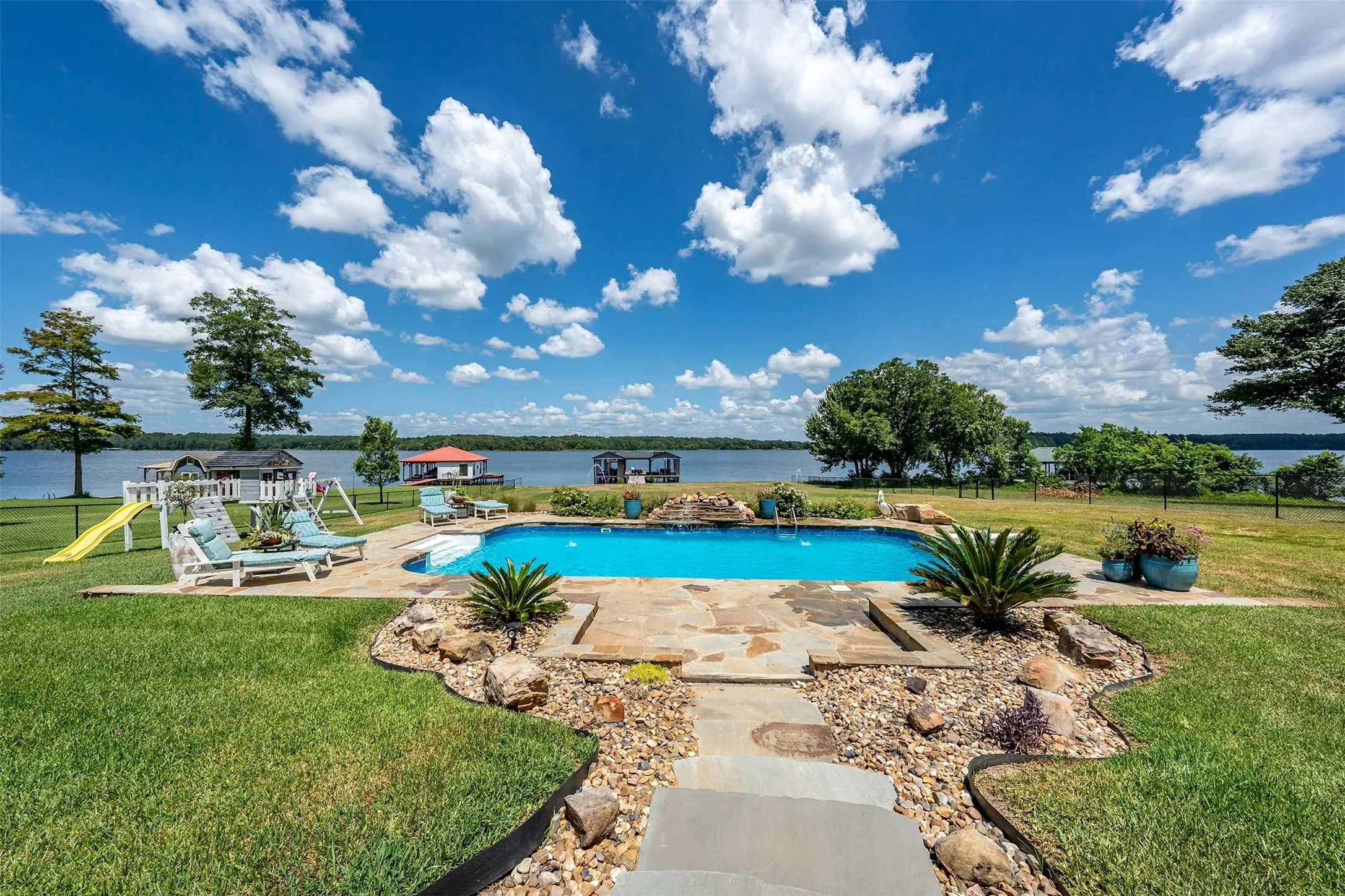 Lakeside property featuring an in-ground swimming pool with stone surround, a stone pathway, and a grassy lawn