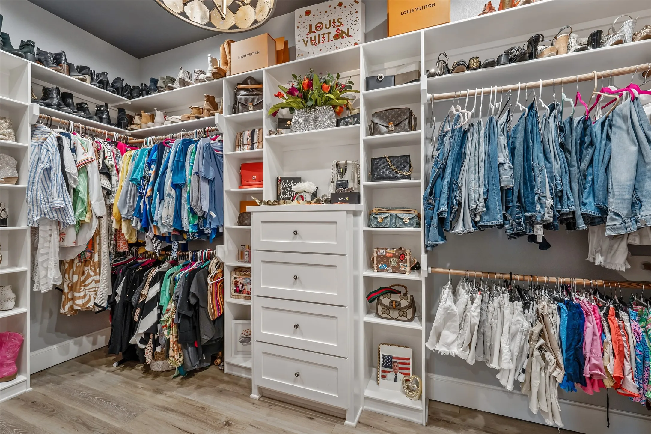 Custom closet system featuring extensive shelving, multiple hanging rods, built-in drawers with dark hardware, and wood-finish flooring