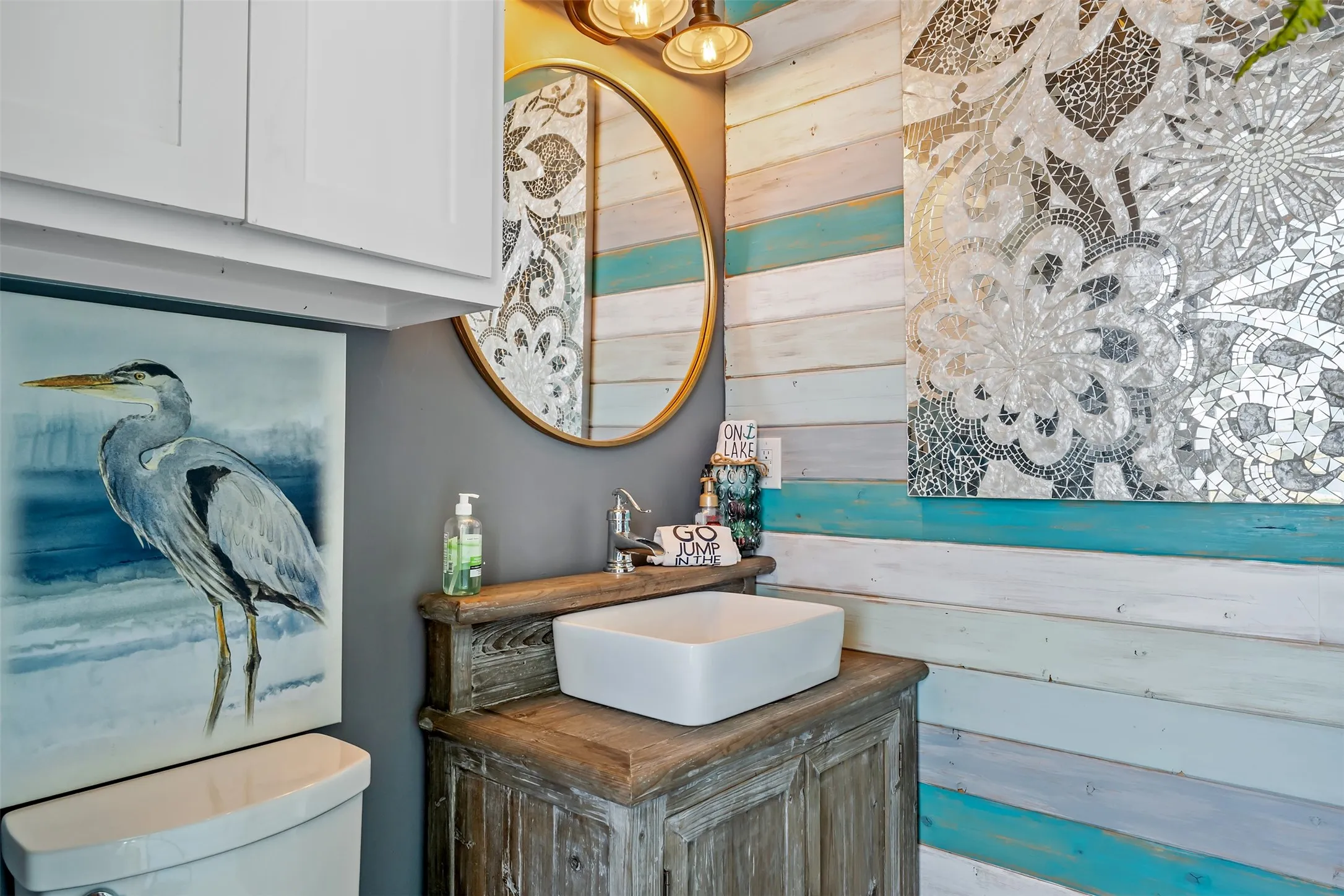 Distressed wood vanity with a white vessel sink, gold-framed round mirror, and a wall-mounted light fixture