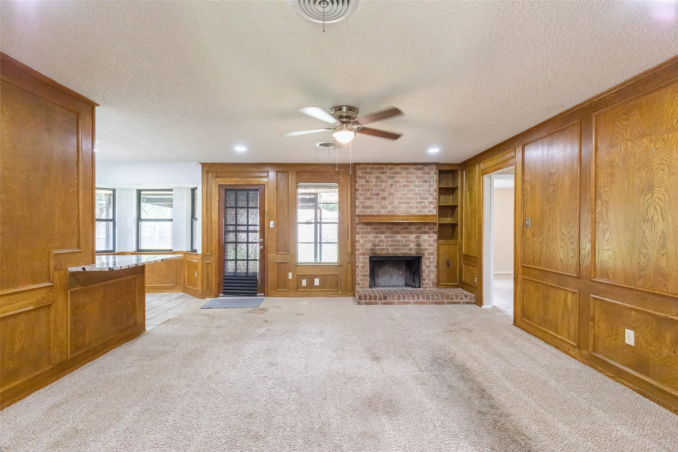 Living area featuring wood paneling, a brick fireplace, built-in shelving, carpet flooring, and a ceiling fan