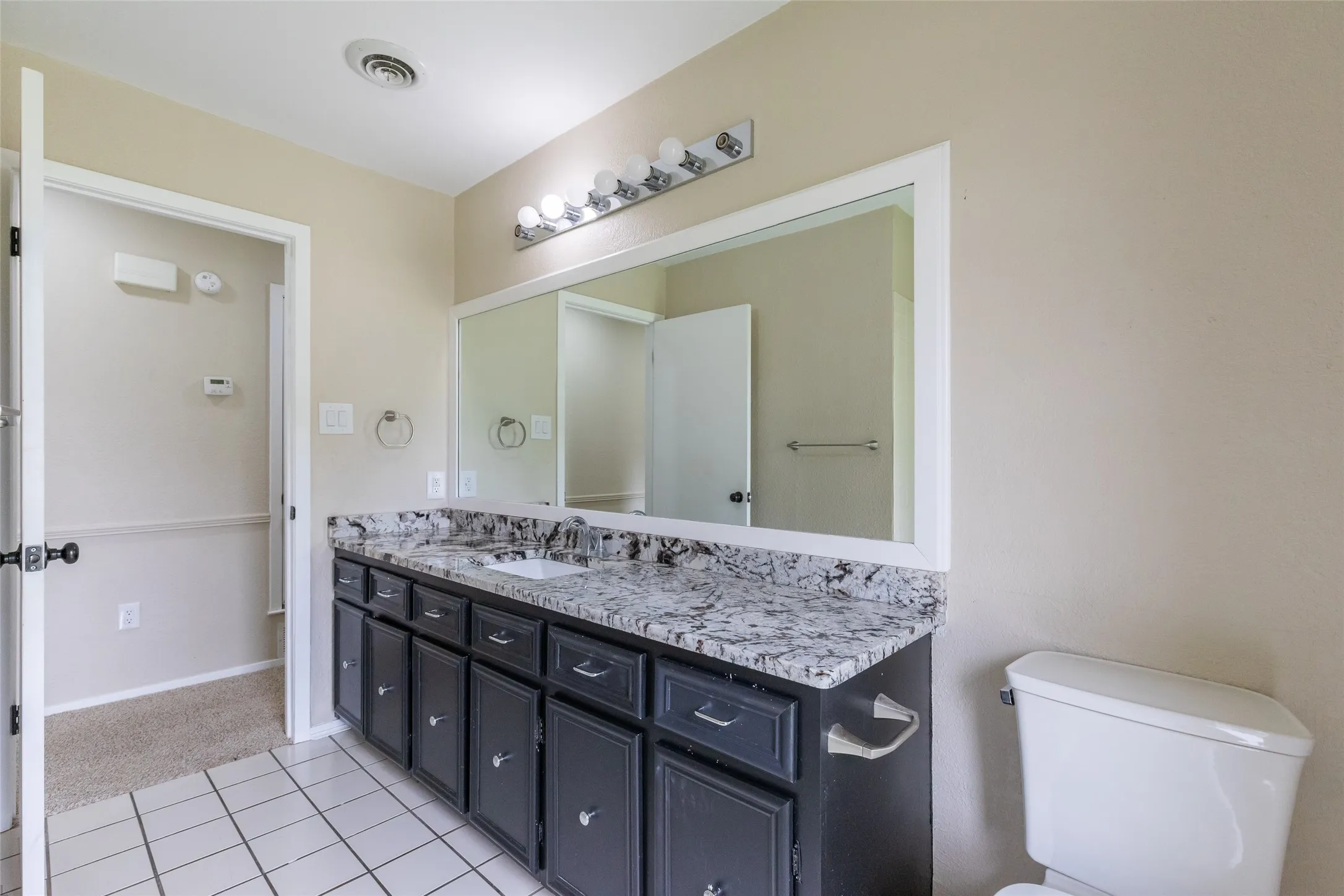 Bathroom featuring a dual vanity with a patterned countertop, white square tile flooring, and a large wall-mounted mirror with overhead lighting