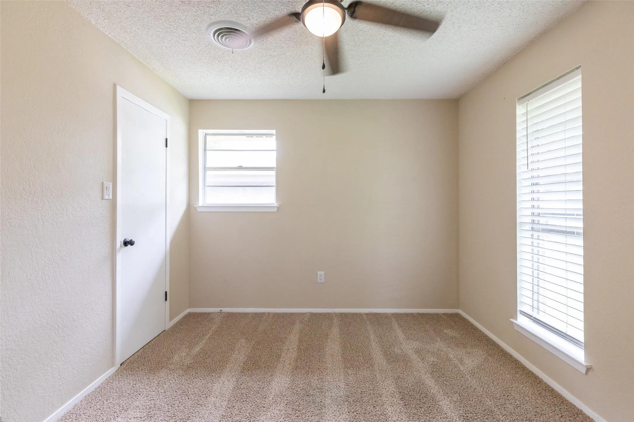 Neutral-toned room featuring a ceiling fan with light fixture, two windows with horizontal blinds, a white interior door with black hardware, and textured light-beige walls