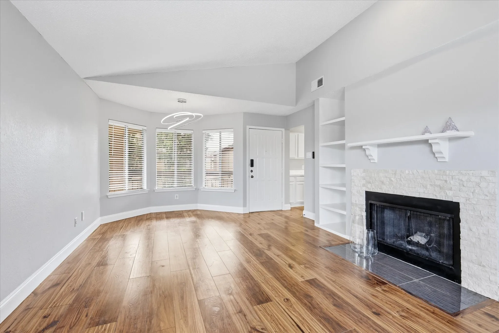 Spacious living area featuring wood-finish flooring, a fireplace with a stone surround, built-in shelving, a modern ceiling light fixture, and multiple windows with blinds