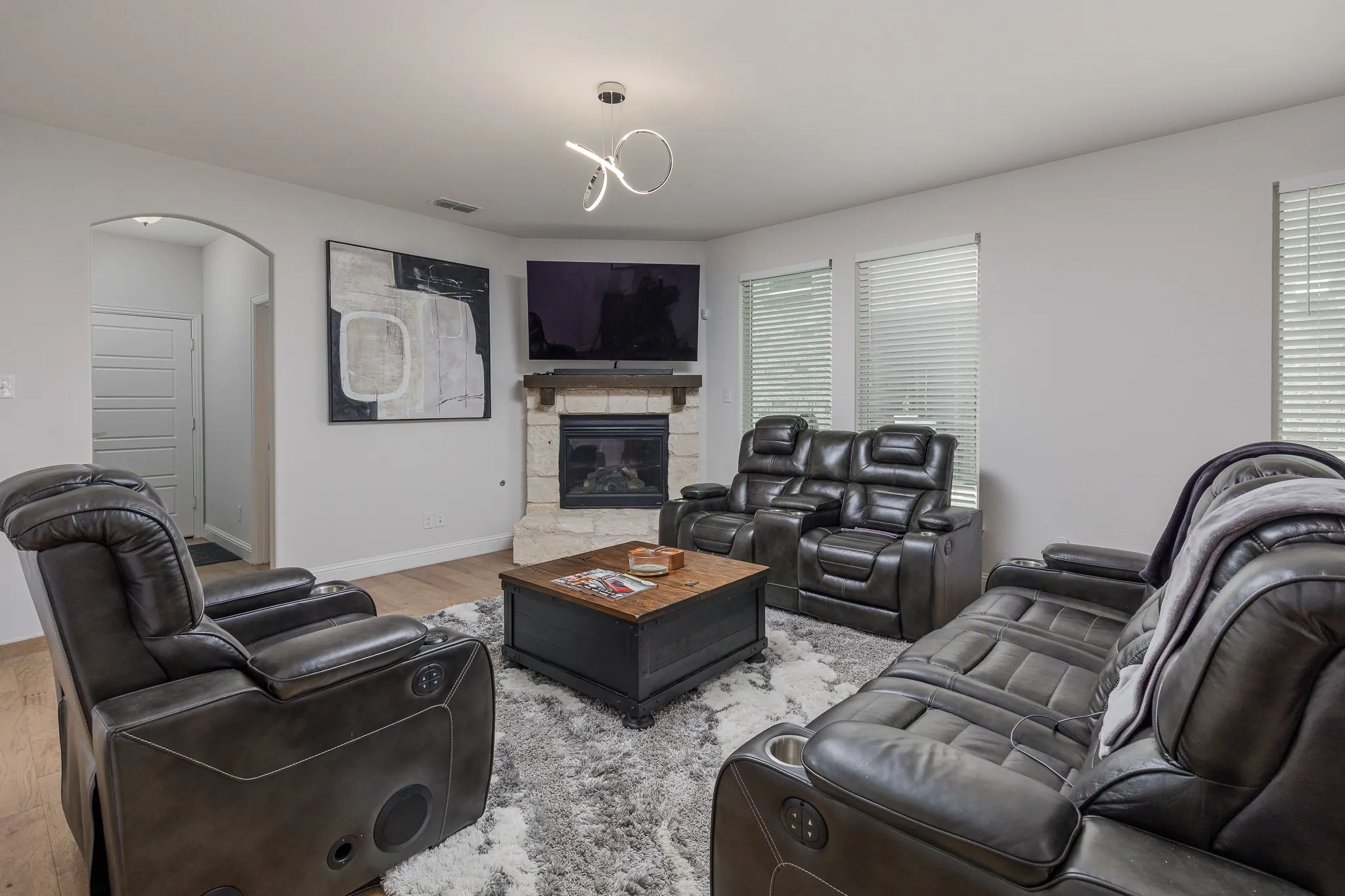 Living area featuring a stone fireplace with a wood mantle, multiple windows with blinds, a contemporary ceiling light fixture, and wood-finish flooring