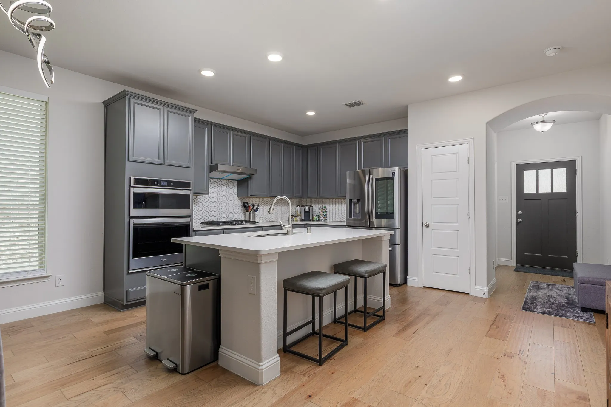 Modern kitchen featuring light wood-finish flooring, grey cabinetry, stainless steel appliances, a white island with seating, and recessed lighting