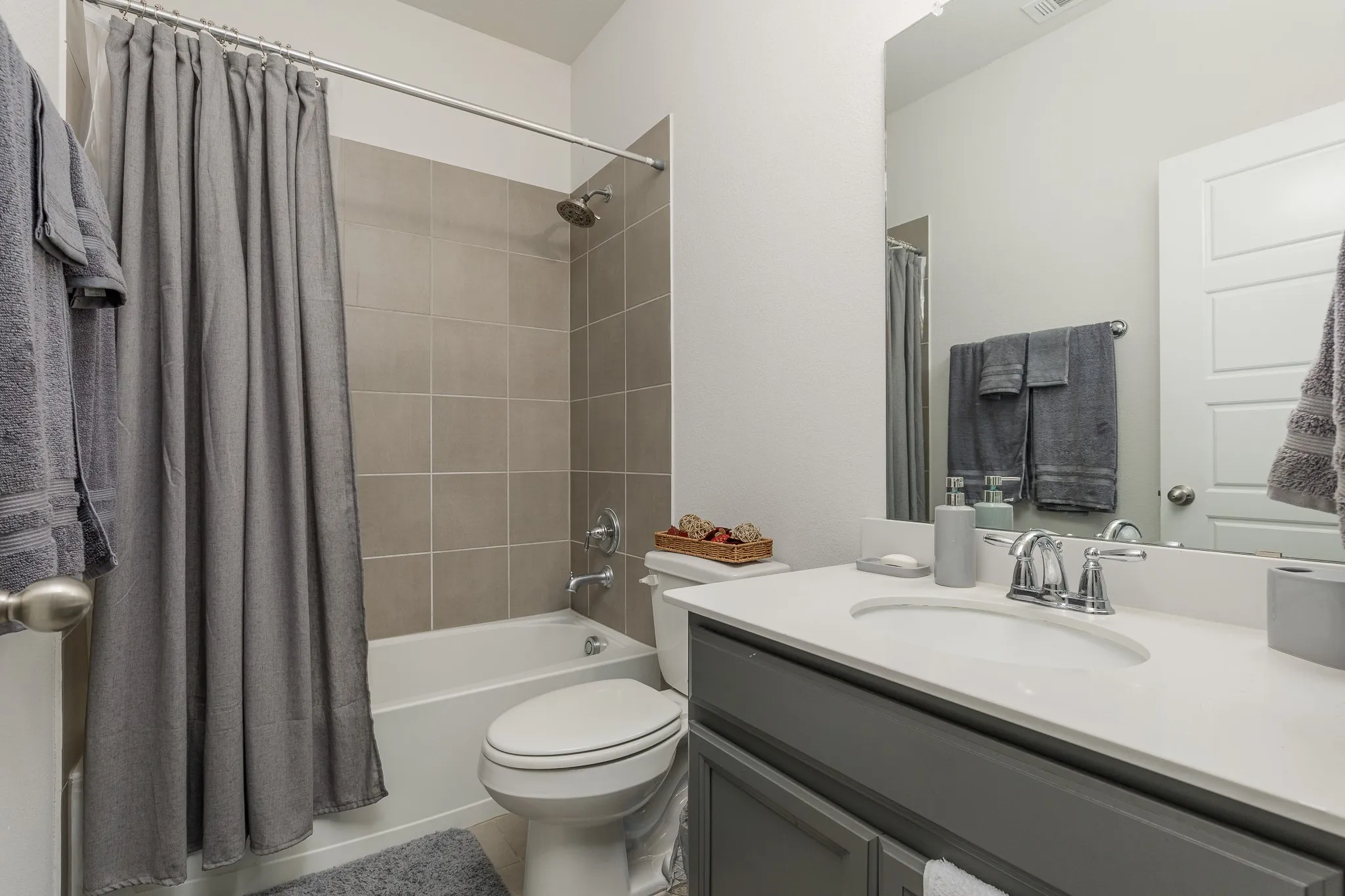 Bathroom featuring a white bathtub, light-toned tiled shower surround, commode, and a vanity with a white countertop and gray cabinetry