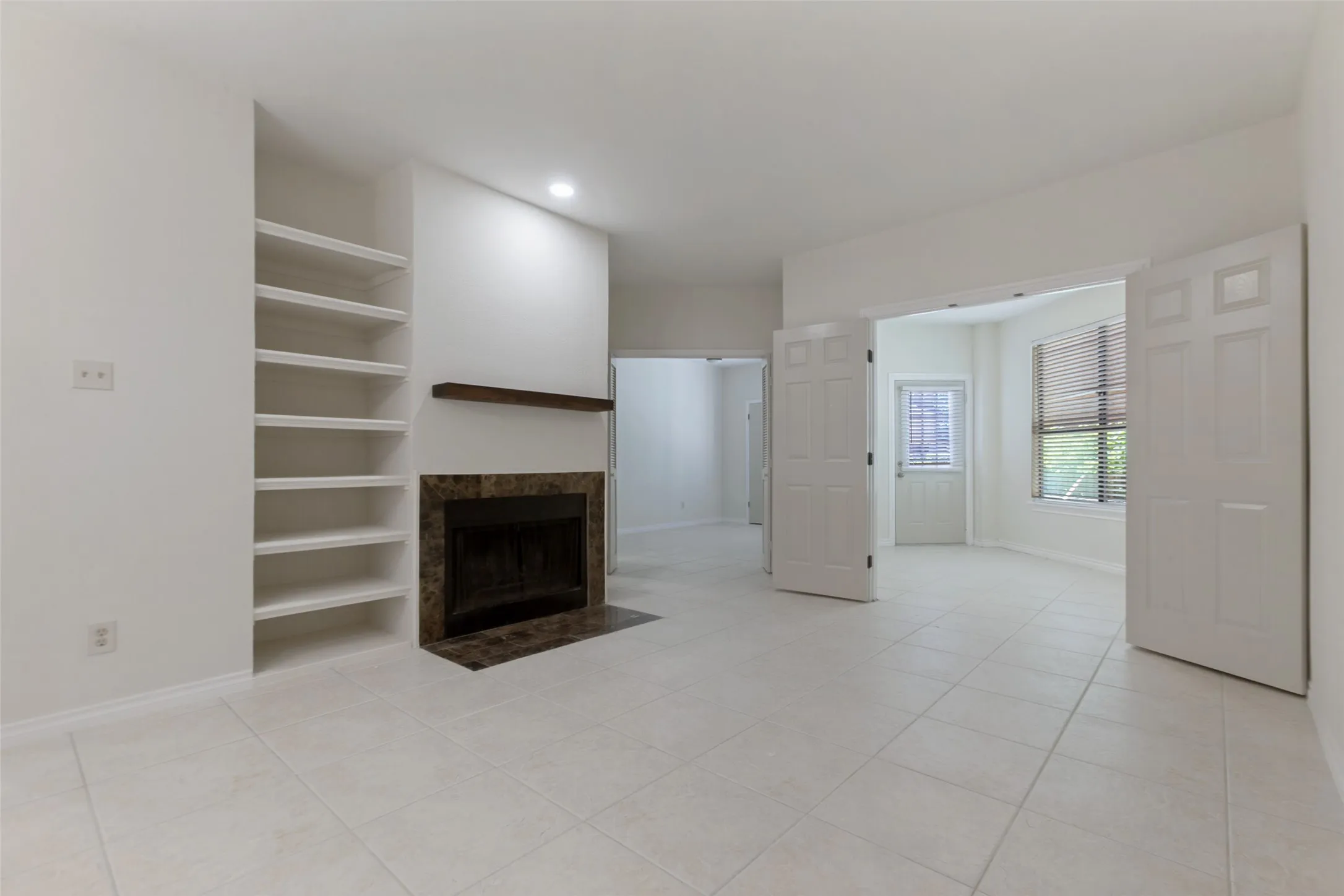 Living area featuring light-toned tile flooring, built-in shelving, a fireplace with a stone surround and wood mantel, and double doors opening to an additional room with a large window
