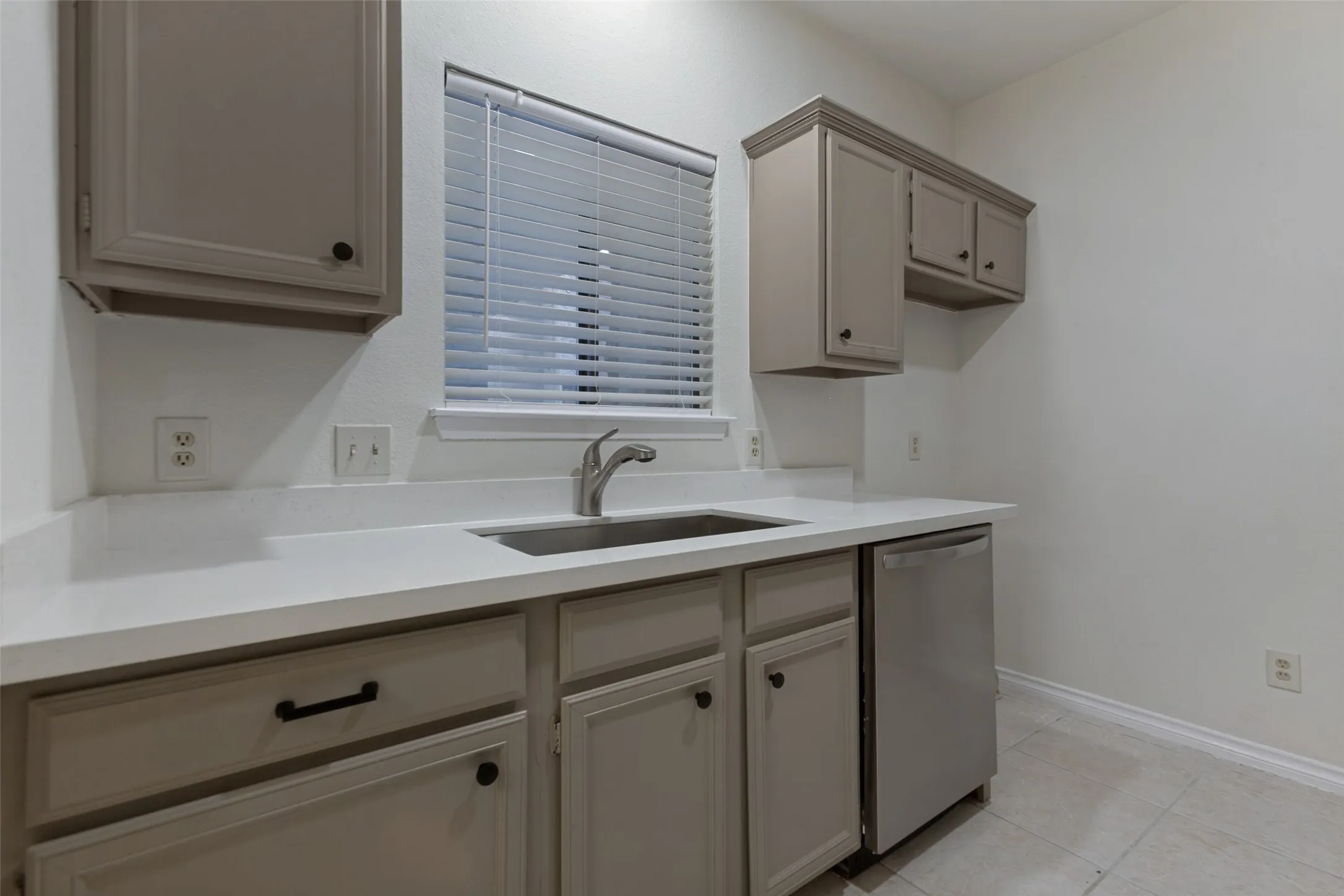 Kitchenette featuring neutral-toned cabinetry, a stainless steel sink, a stainless steel dishwasher, white countertops, and tile flooring