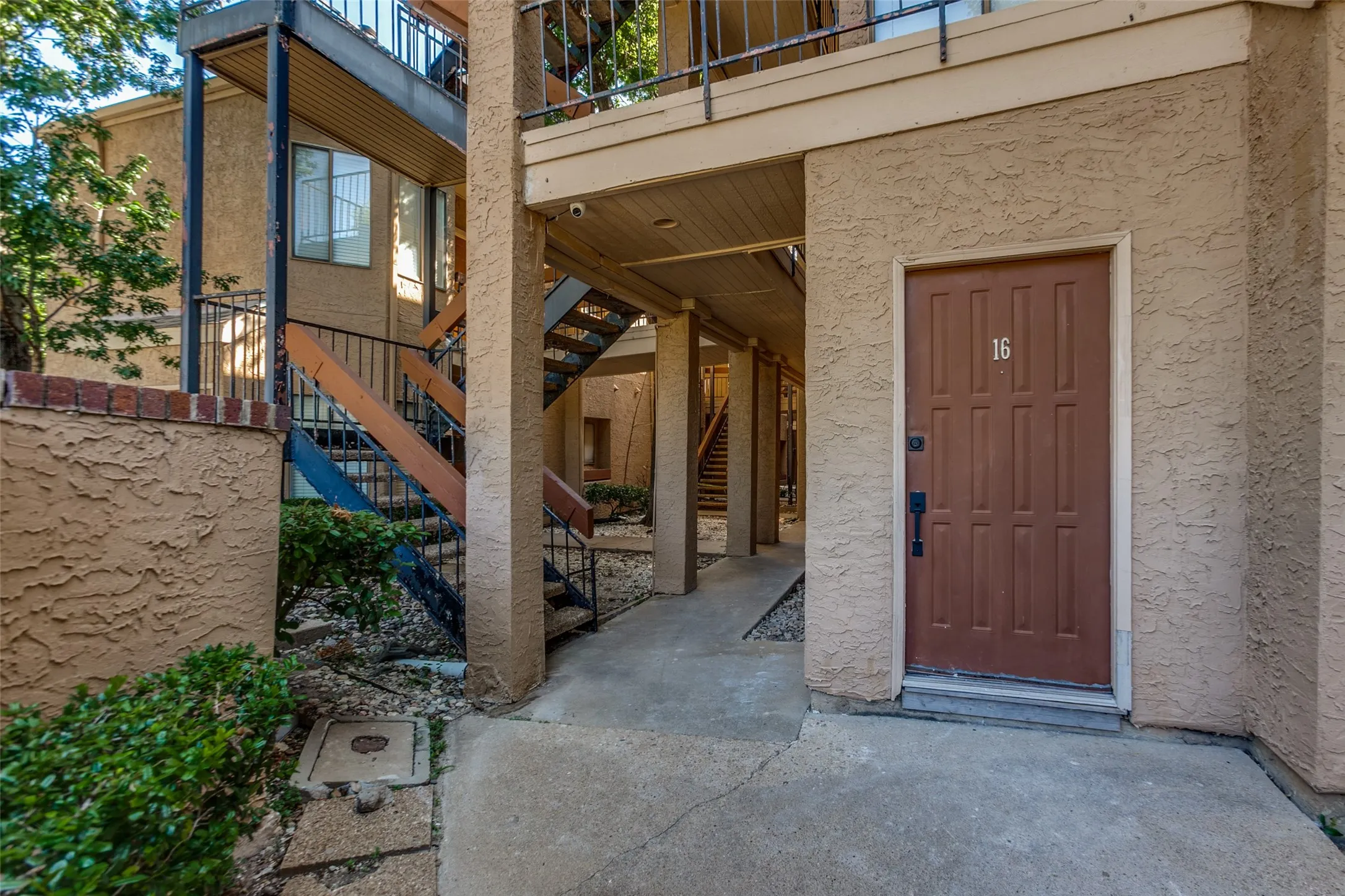 Stucco exterior entryway featuring a paneled door with numerical address, concrete pathway, and mature landscaping