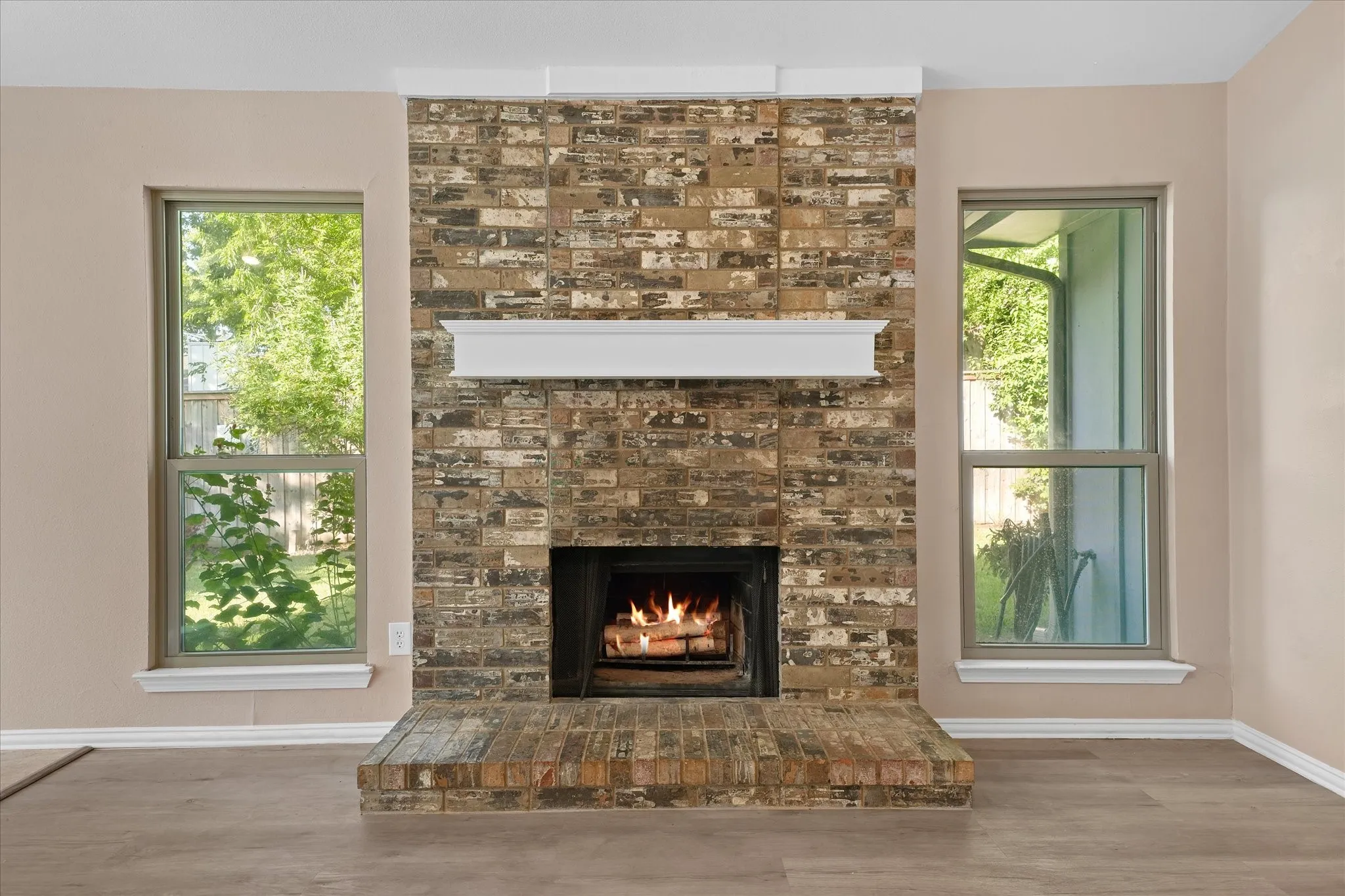 Brick-face woodburning fireplace with a white mantel, flanked by two single-hung windows, and wood-finish LVP flooring