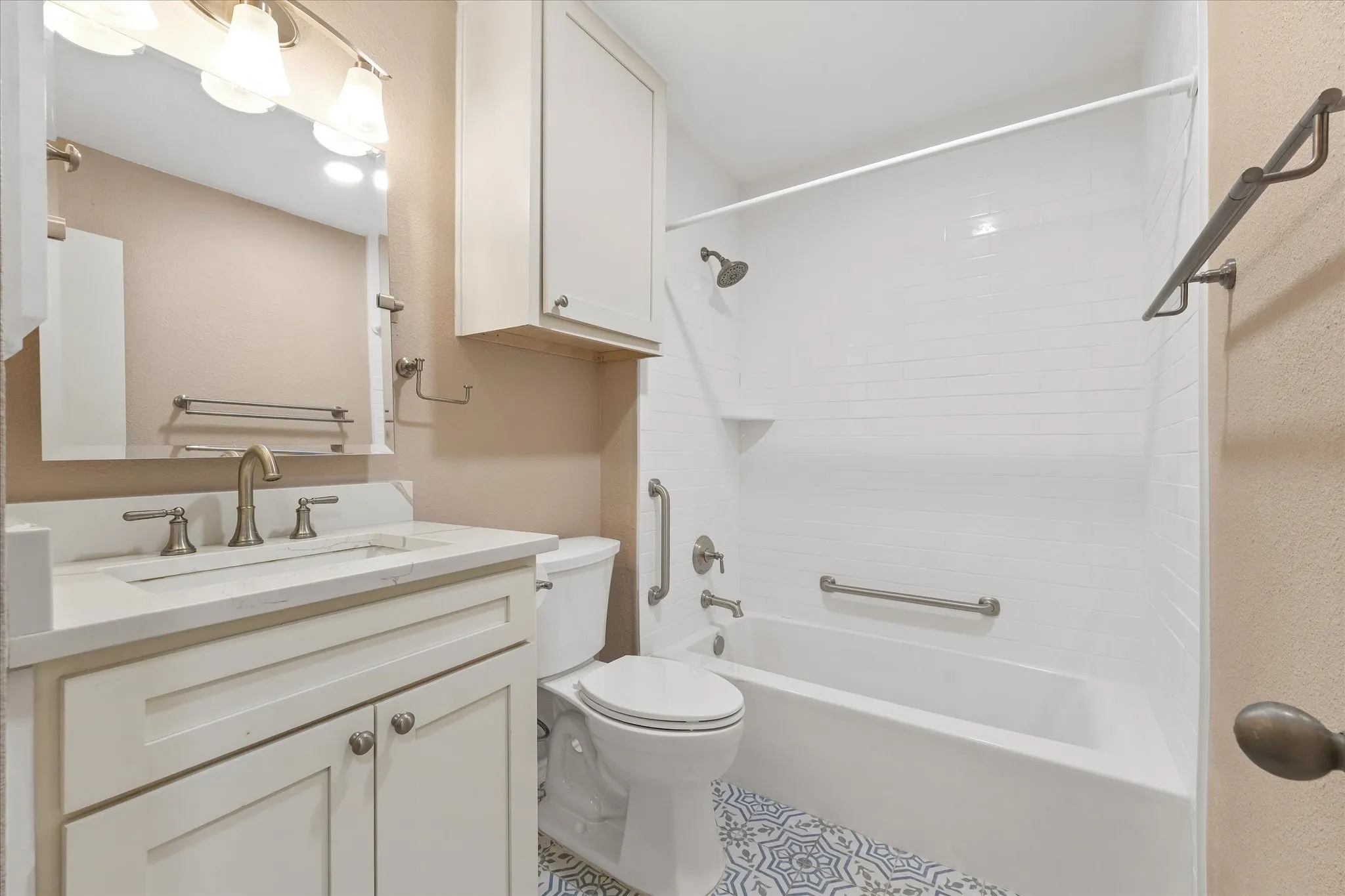 White vanity with an integrated sink and brushed nickel faucet and quartz counter, matching overhead cabinetry, white subway-tiled shower, and decorative floor tile