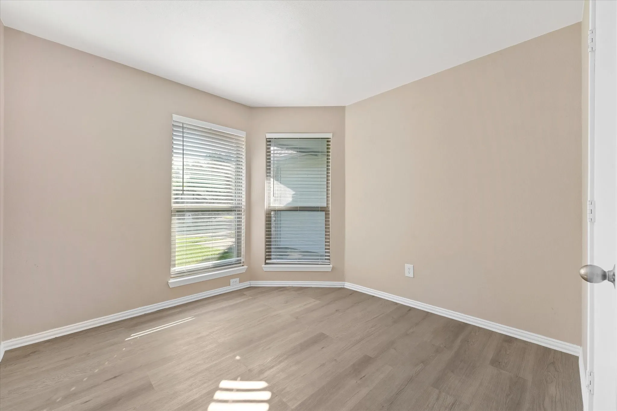 Light-toned walls with white trim, wood-finish LVP flooring, two large windows with blinds, and a white door with a brushed nickel knob