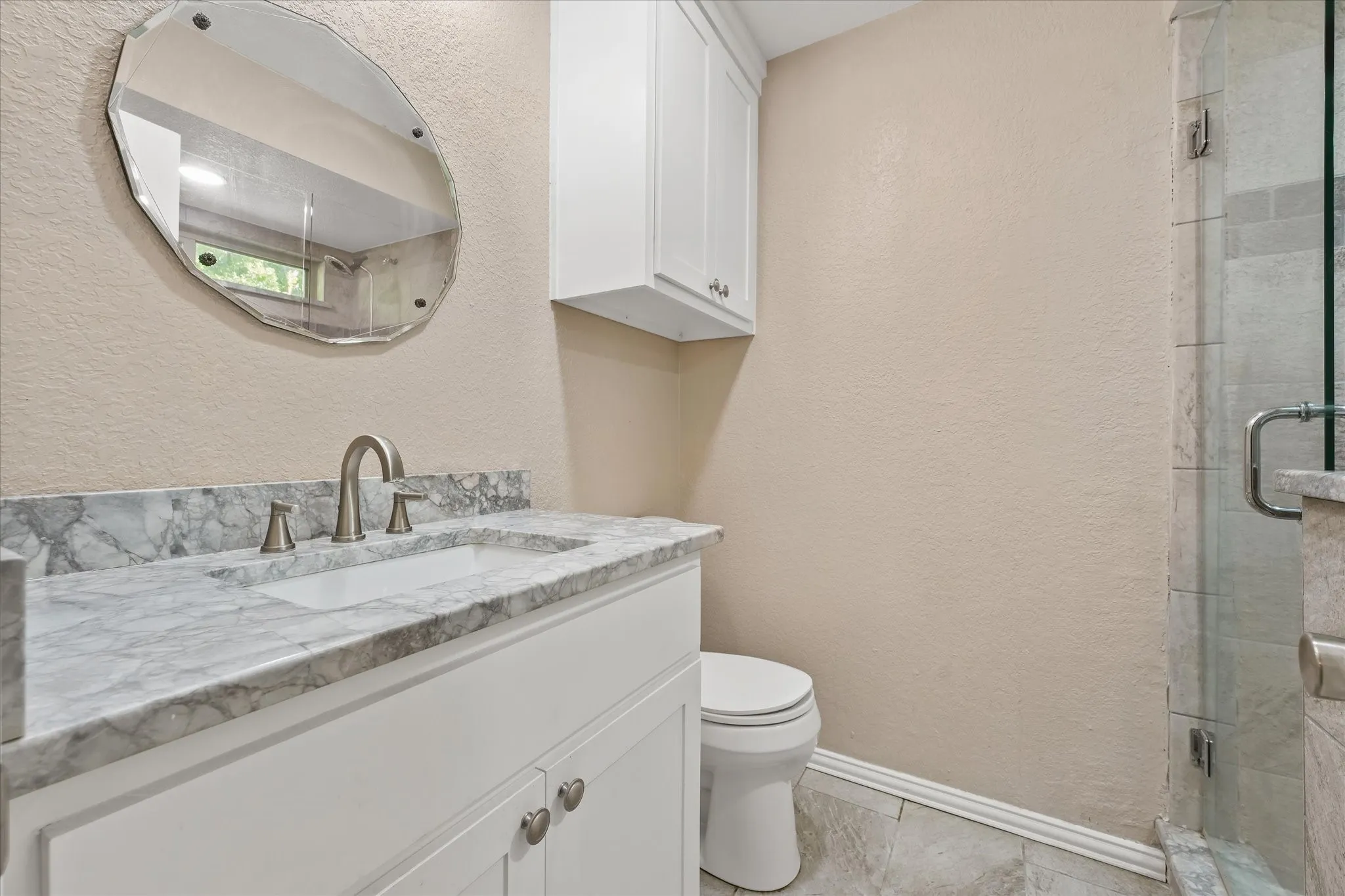 White vanity with a quartz countertop and integrated sink, brushed nickel faucet, wall-mounted storage cabinet, framed mirror and updated tile shower.