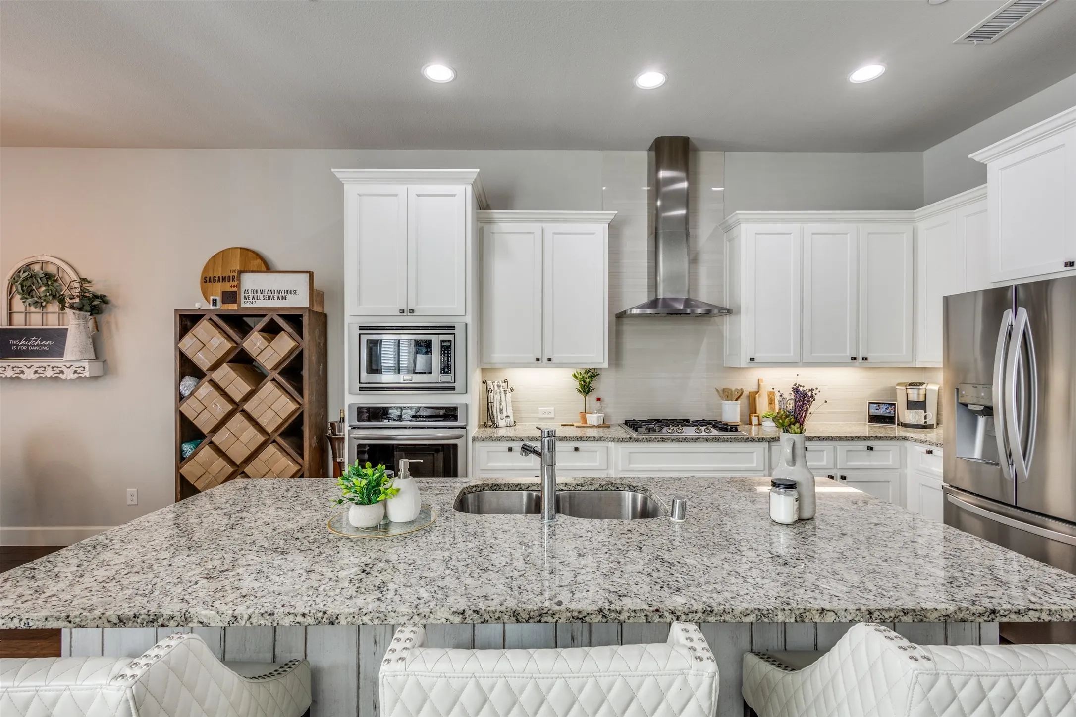 Spacious kitchen featuring an oversized island with double sink, white cabinetry, integrated stainless steel appliances, stainless steel range hood, and recessed lighting
