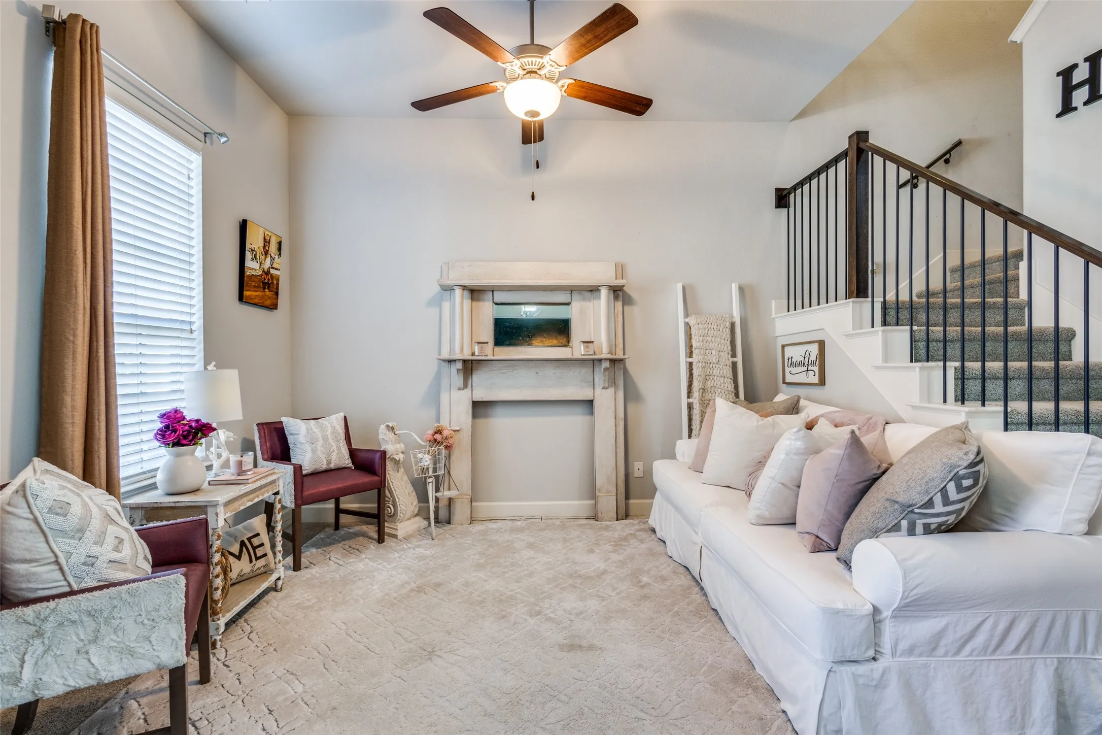 Inviting living area featuring a decorative mantel, wood-finish ceiling fan, window with blinds, carpeted flooring, and a staircase with dark wood-finish handrails and black metal balusters