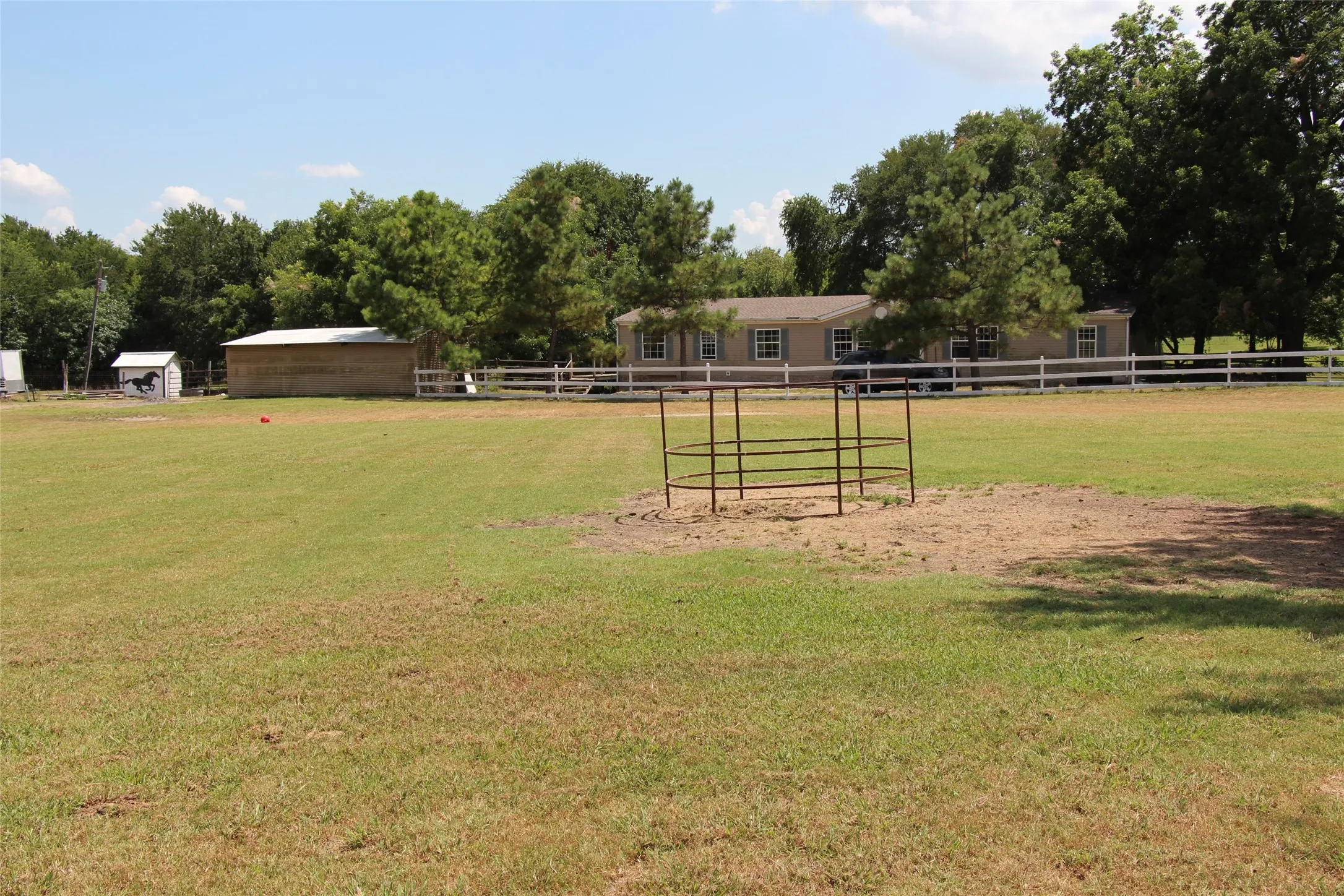 Expansive green space featuring a white perimeter fence, hay feeder, mature trees, a detached storage building, and a tan single-story dwelling with white-trimmed windows