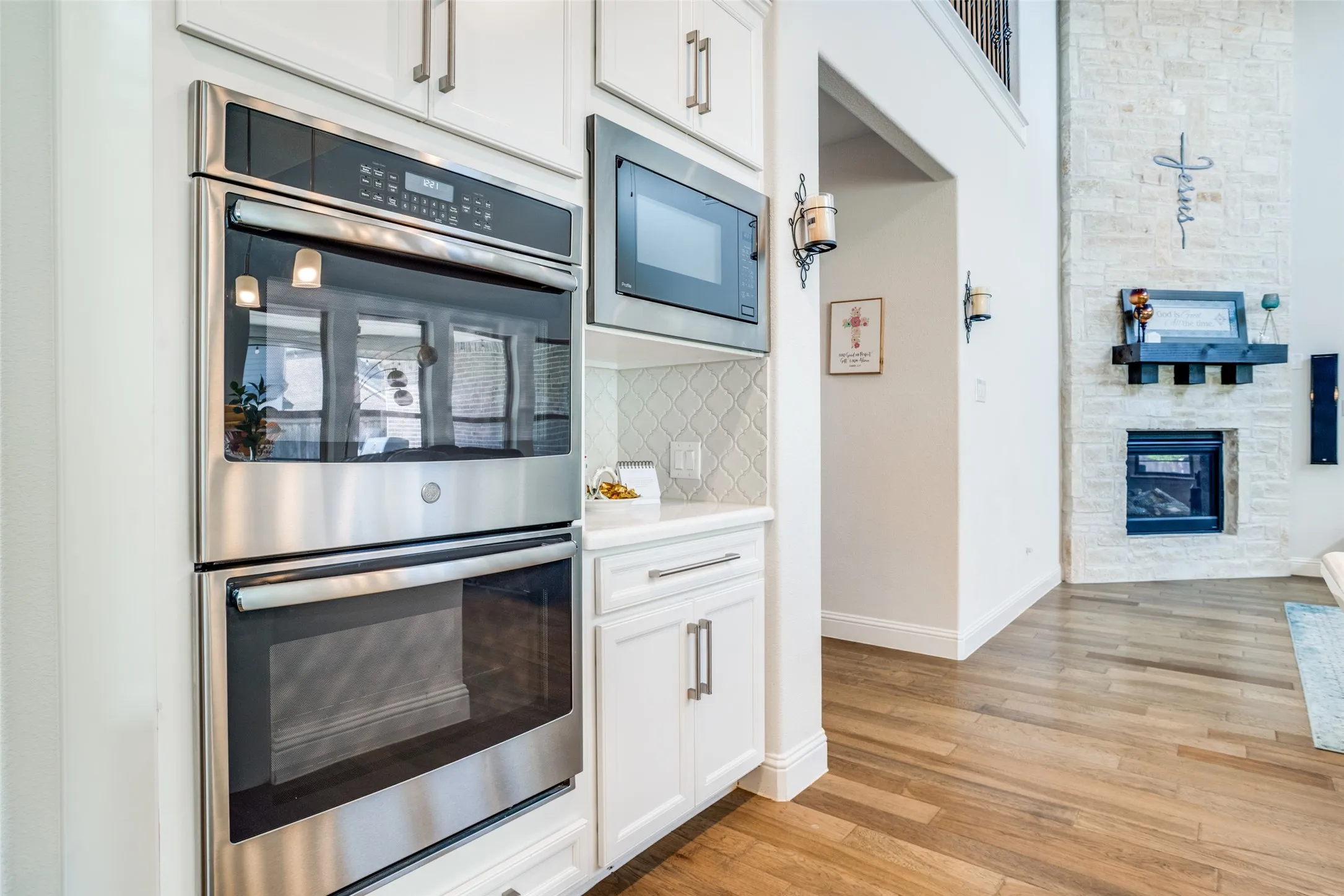 Stainless steel double wall ovens and microwave, white cabinetry with brushed nickel hardware, diamond pattern tile backsplash, wood-finish flooring, and a floor-to-ceiling stone fireplace
