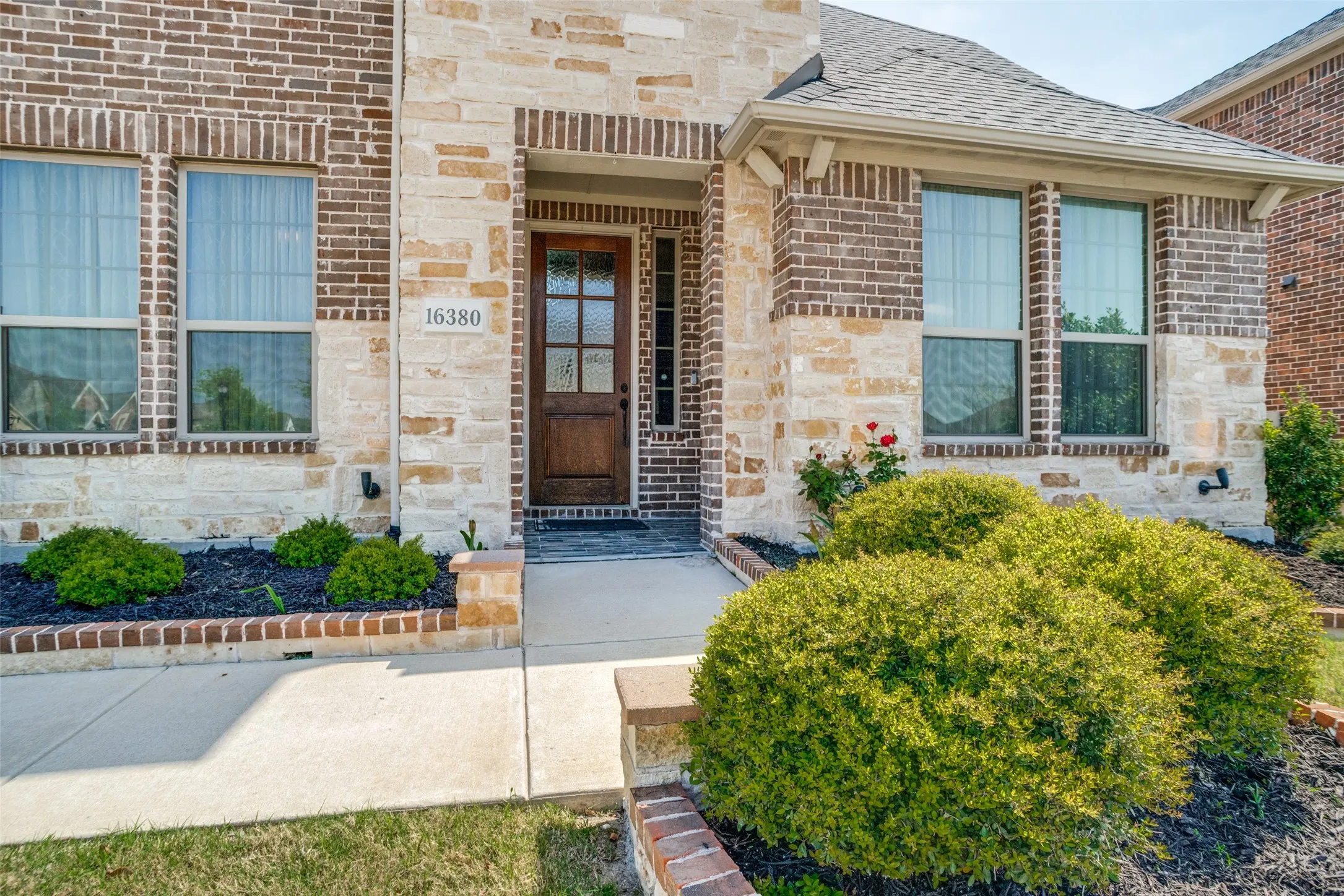 Exterior facade featuring brick and stone construction, a paneled wood front door with glass inserts, mature landscaping, and a concrete pathway leading to the entrance