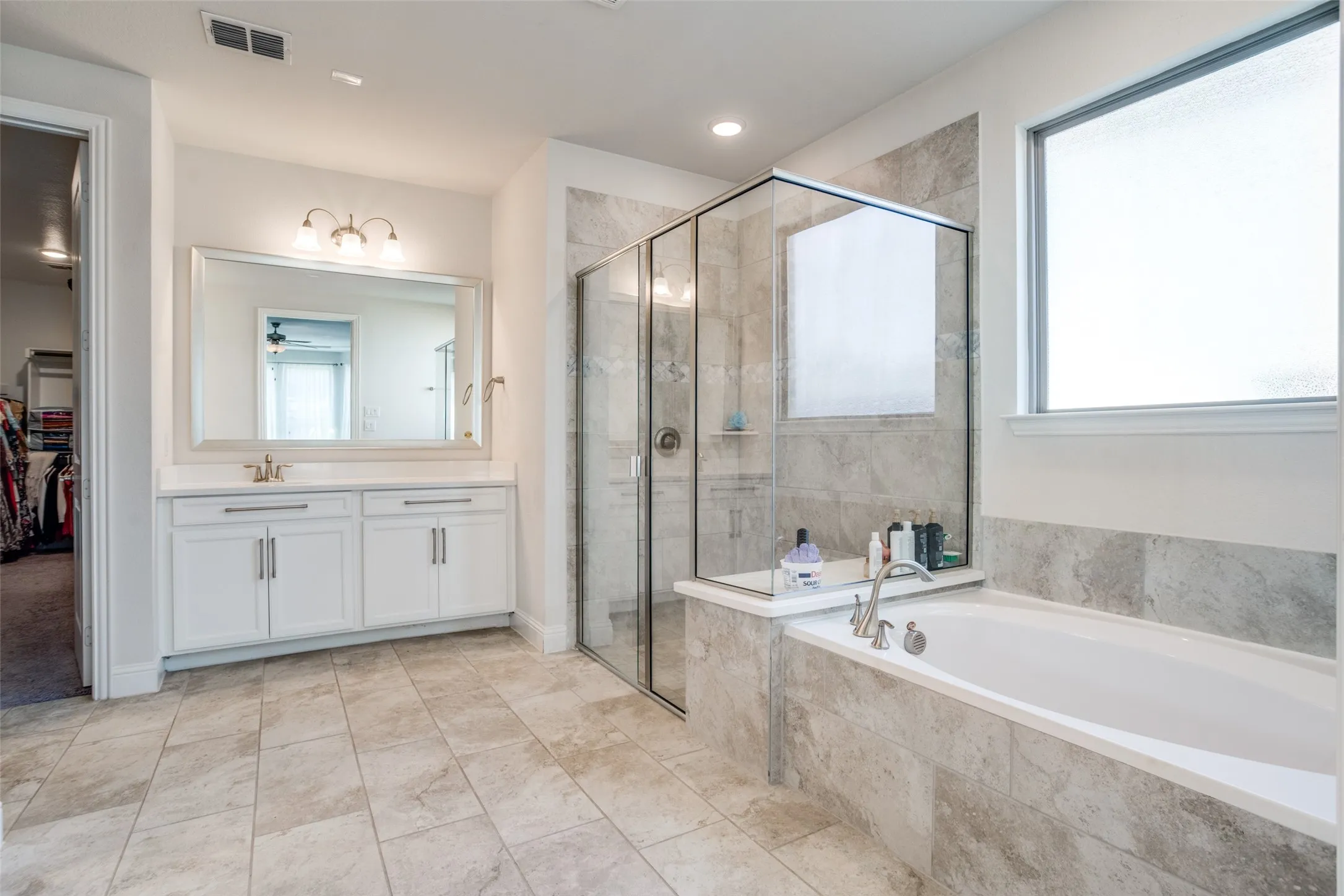 Spacious bathroom featuring a built-in soaking tub, a frameless glass shower enclosure, a dual-sink vanity with white cabinetry and a large mirror, and ceramic tile flooring