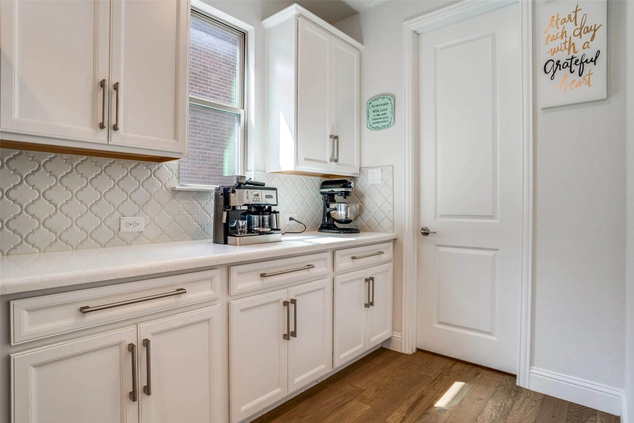 White shaker cabinetry with brushed nickel hardware, light-toned countertop, quatrefoil tile backsplash, and wood-finish flooring