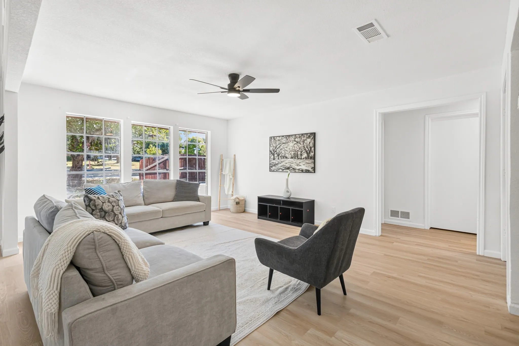 Bright and spacious living area showcasing gleaming wood-finish flooring, abundant natural light from multi-pane windows, a ceiling fan with dark finish, neutral wall tones, and a black media console with open shelving
