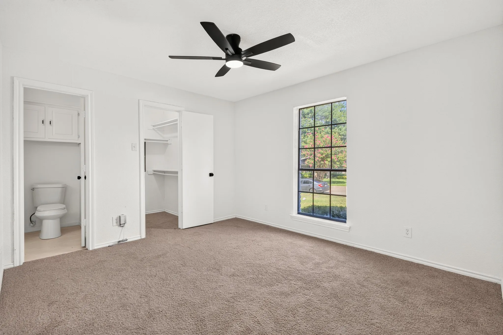 Carpeted room featuring a ceiling fan, a window with black grid panes, built-in shelving, and a toilet with overhead cabinetry