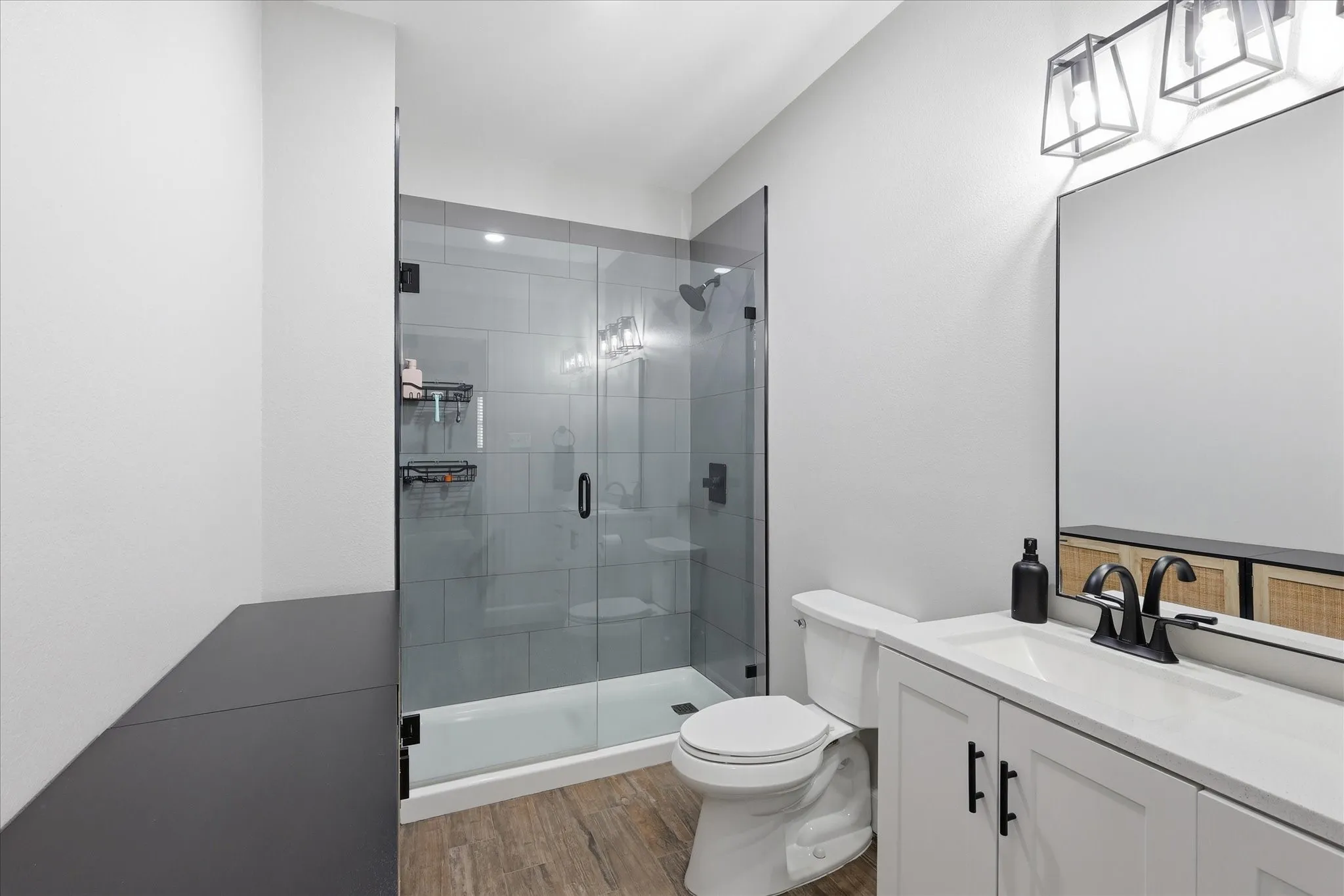 Contemporary bathroom featuring a glass-enclosed shower with tile surround, a white vanity with integrated sink, wood-finish flooring, and black fixtures
