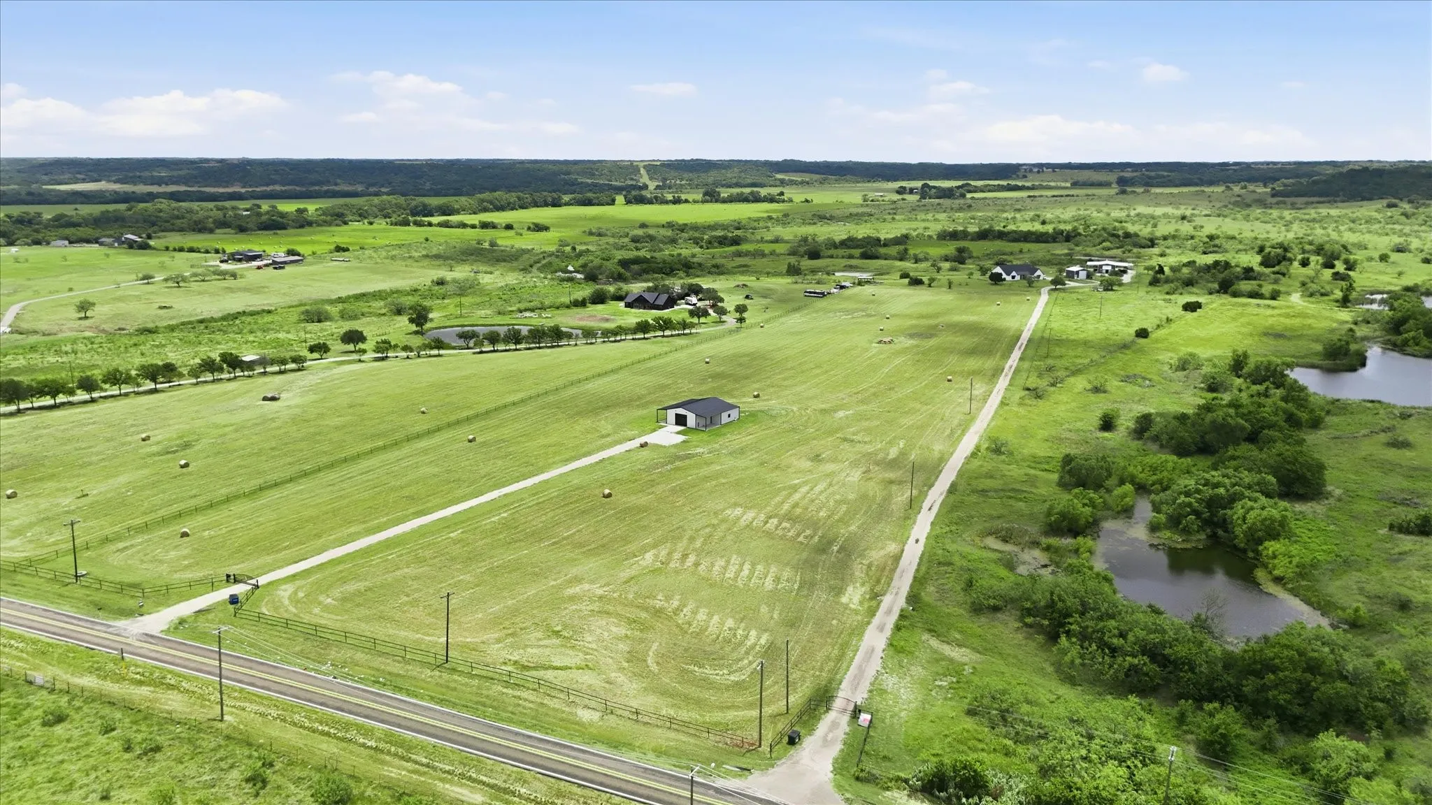 Expansive rural property featuring a paved road, a single building with a dark roof and white siding, a long gravel driveway, and multiple ponds surrounded by lush greenery