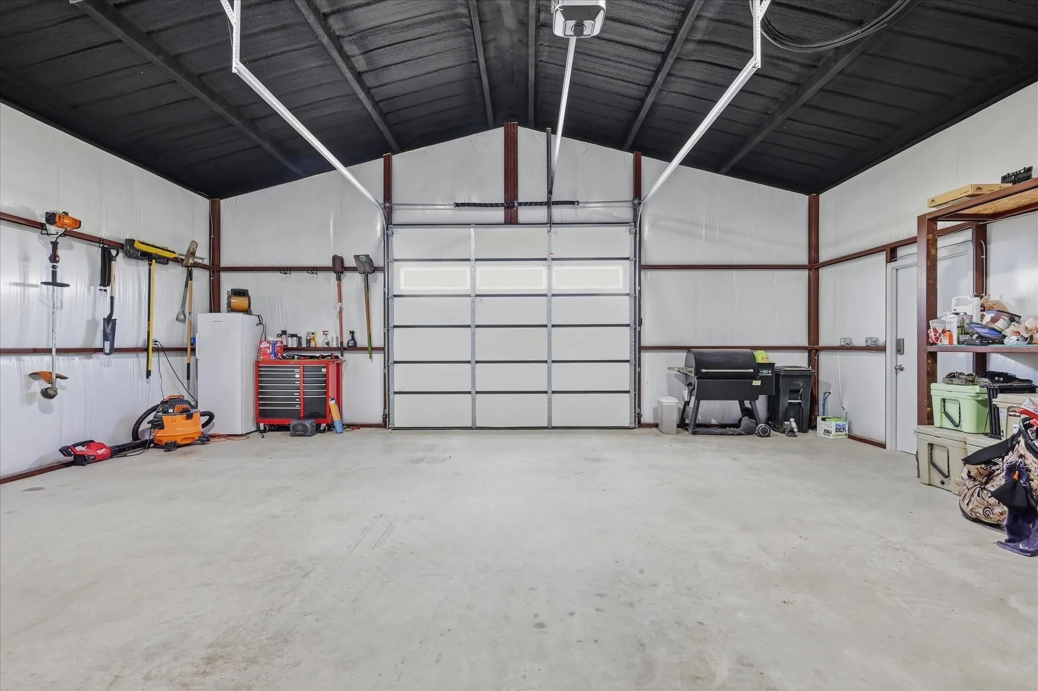Spacious utility structure featuring a large overhead door with window panel, concrete flooring, corrugated metal walls, exposed structural beams, and a separate service door