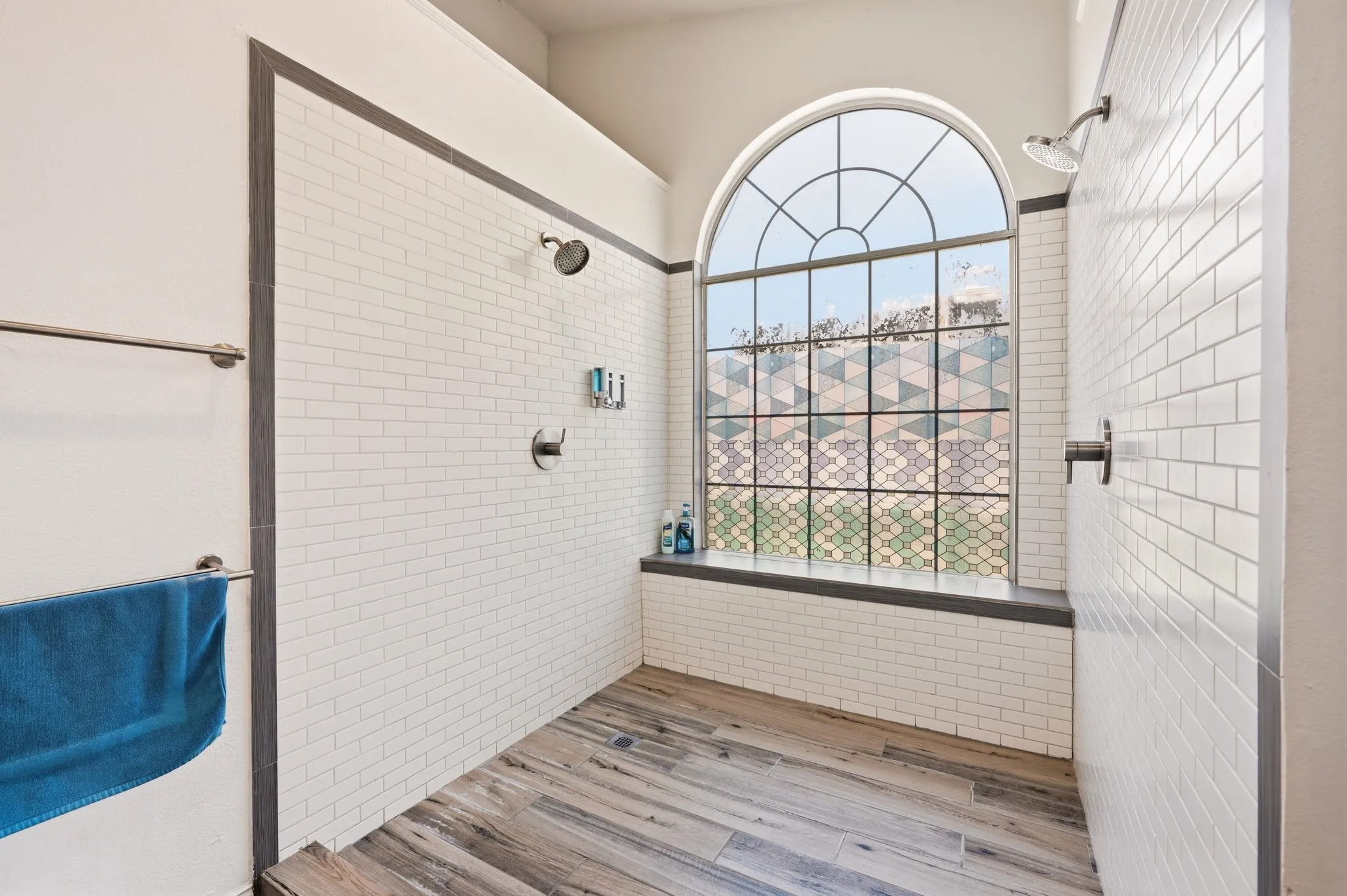 Spacious shower enclosure featuring an arched window, wood-finish flooring, white subway tile walls, and a built-in bench