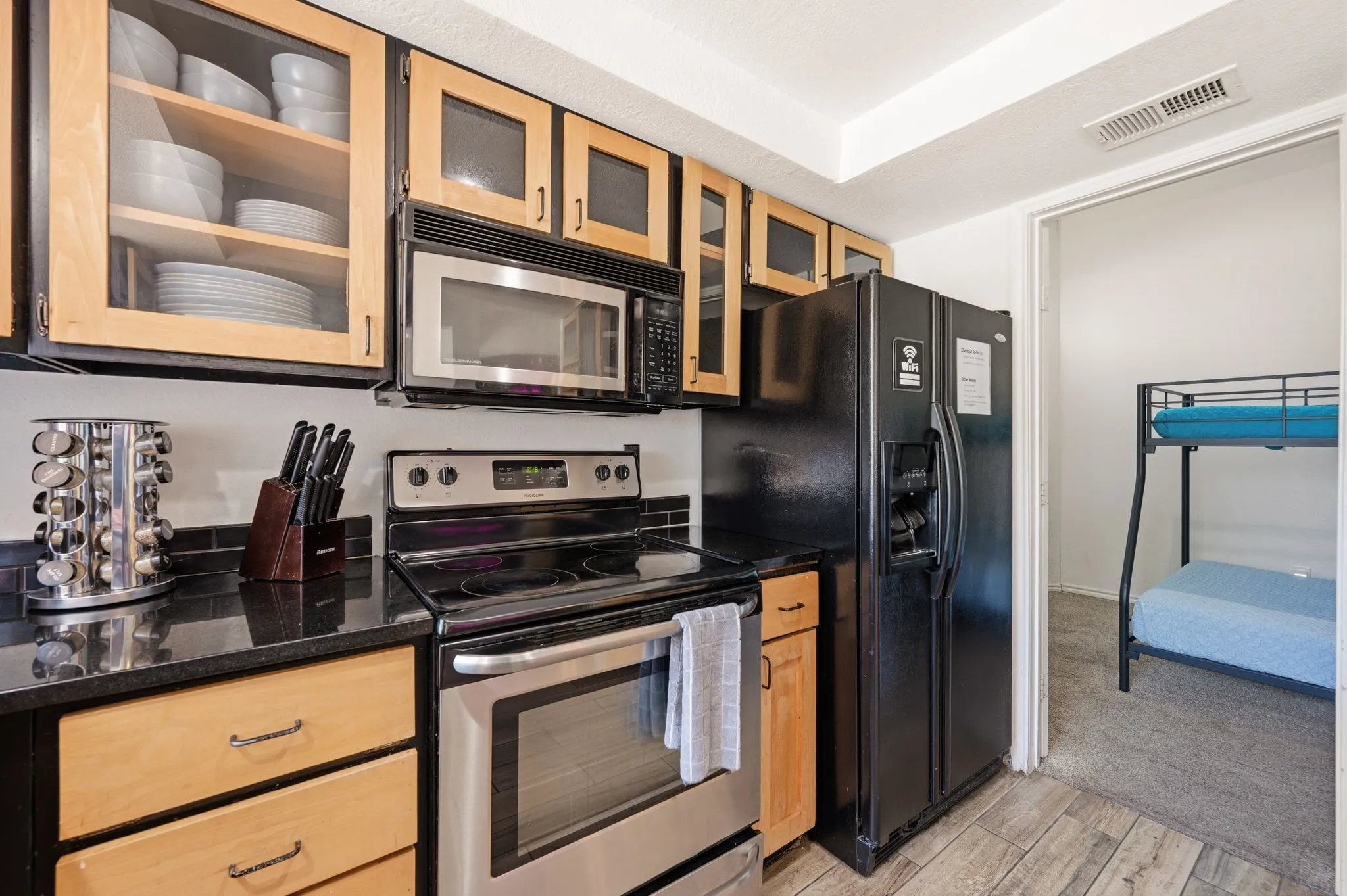Kitchen featuring granite-look countertops, black tile backsplash, wood-finish cabinetry with glass-front uppers, and stainless steel appliances including a microwave and range
