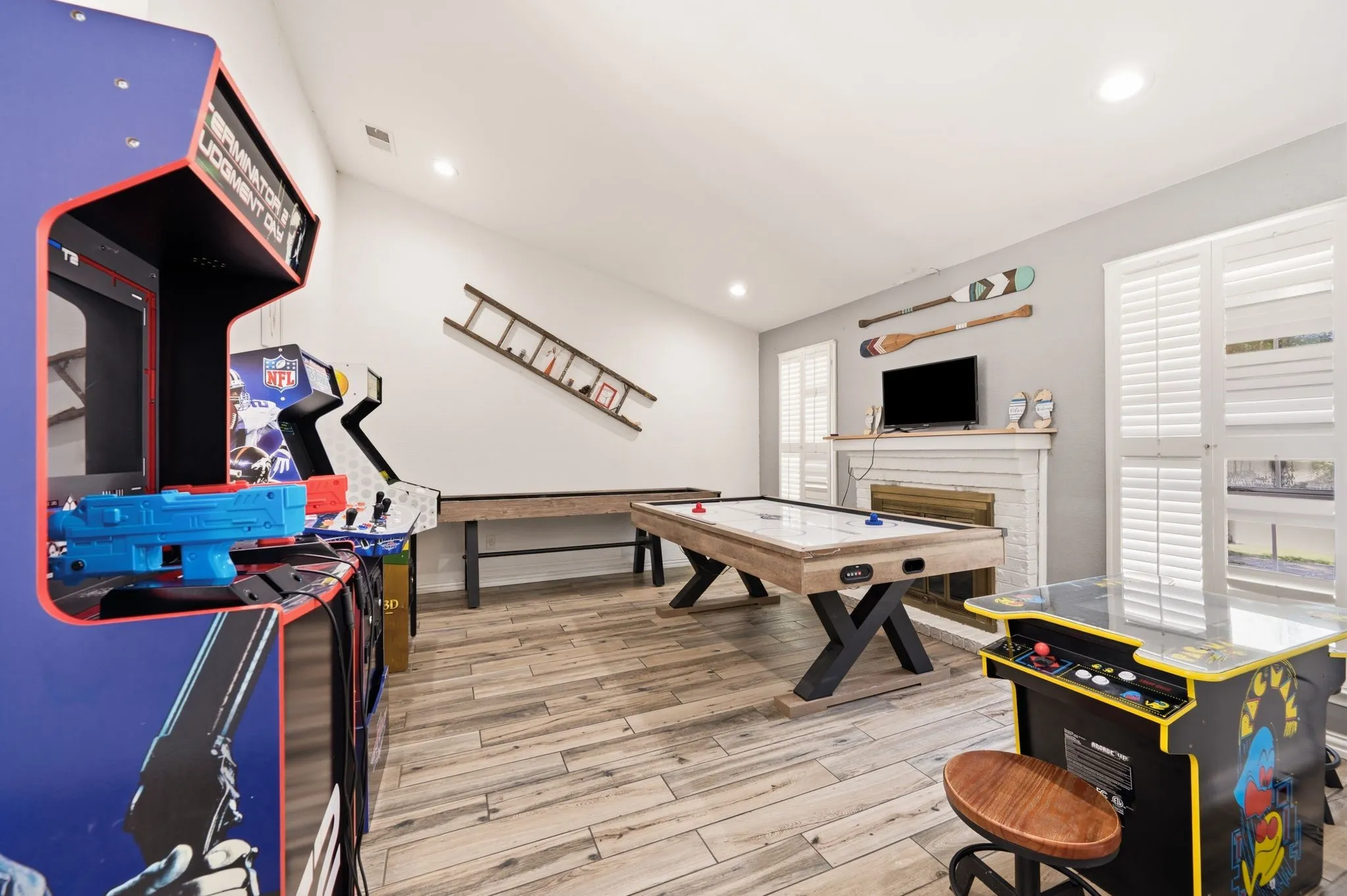 Spacious recreation room featuring wood-finish flooring, a white mantel, and multiple windows with plantation shutters