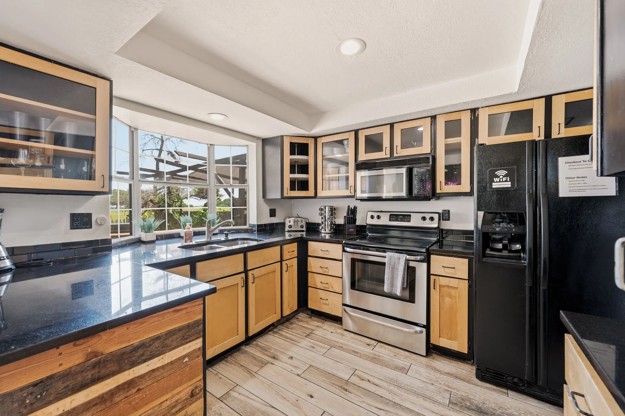 Kitchen featuring light wood cabinetry with glass-front uppers, black countertops, and a bay window over the sink