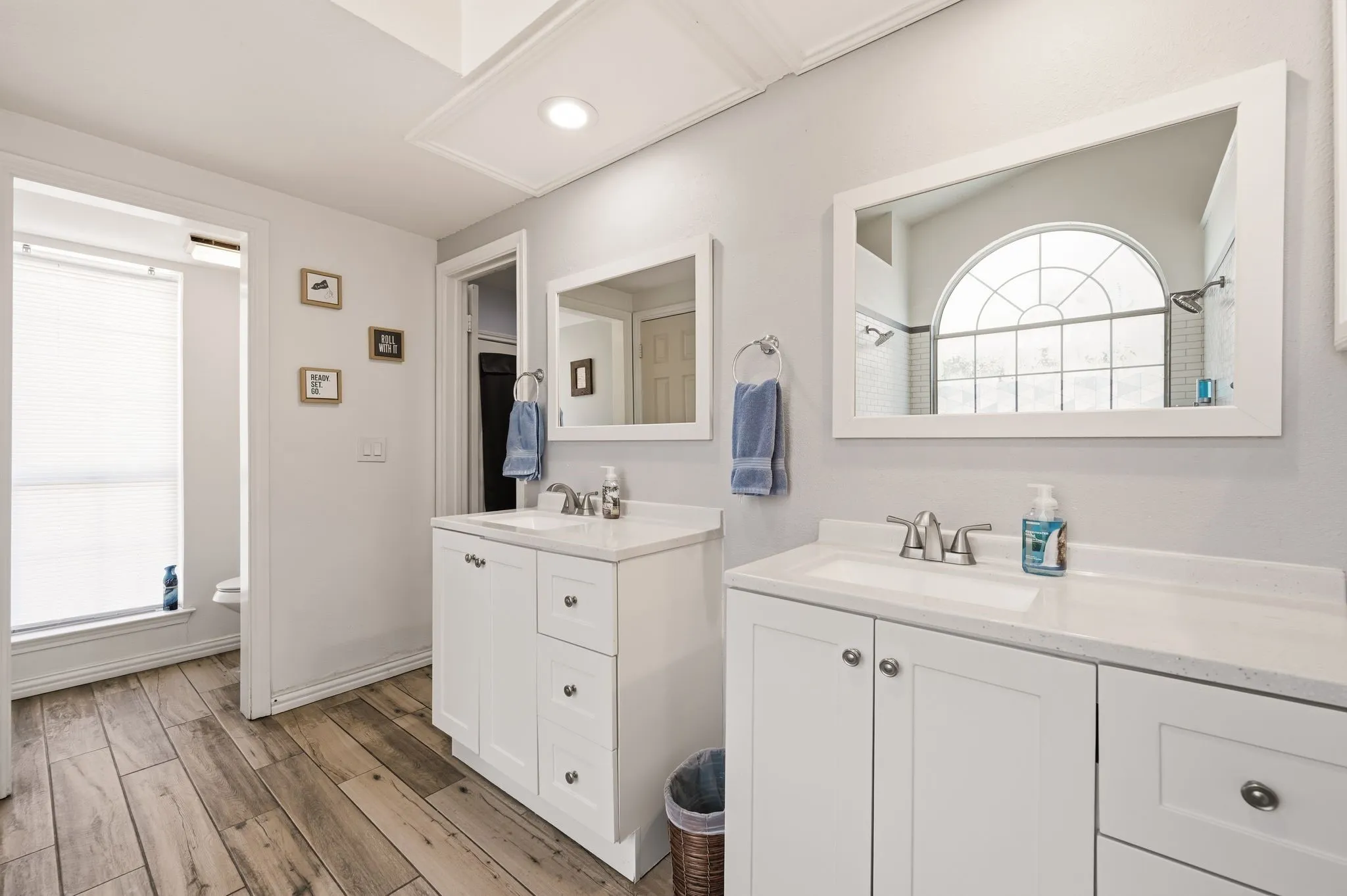 Bathroom featuring dual white vanity cabinets with integrated sinks and white countertops, individual framed mirrors, wood-finish flooring, and a large arched window with grid details