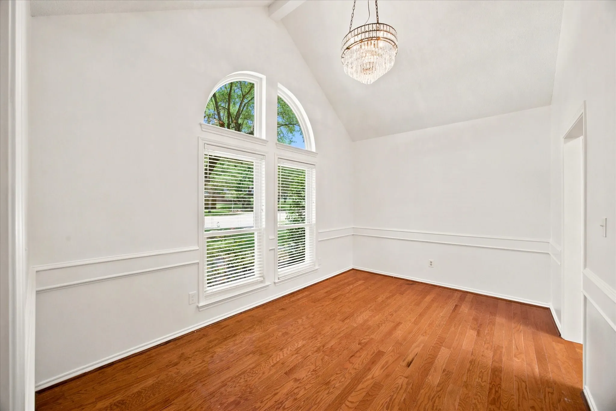 Vaulted ceiling room featuring hardwood flooring, extensive natural light from a large arched window, a crystal chandelier, and wainscoting detailing
